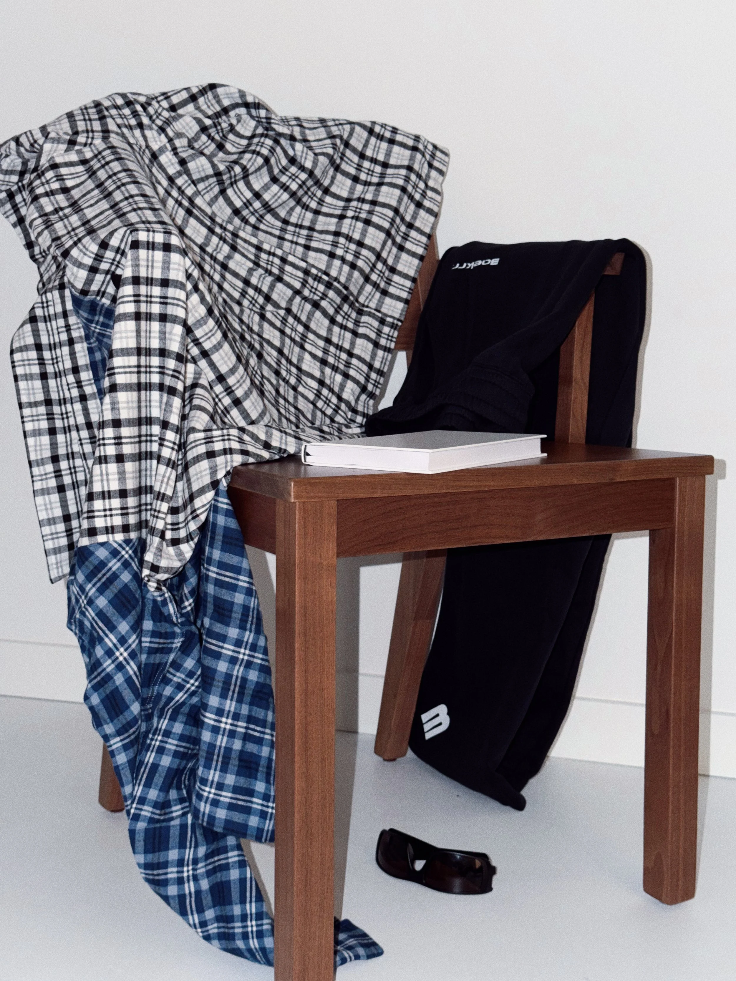A plaid shirt draped over a wooden table with a closed notebook, a black clothing item, and a single sandal on the floor next to the table.