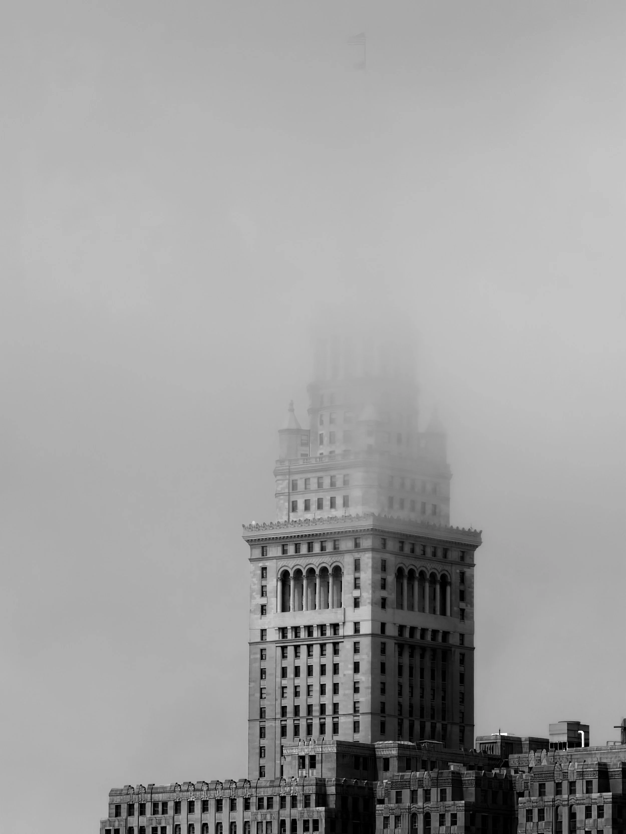 A tall building with a distinctive top, partially covered by fog, is shown in black and white. The building has a classical architectural style with detailed columns and edges.