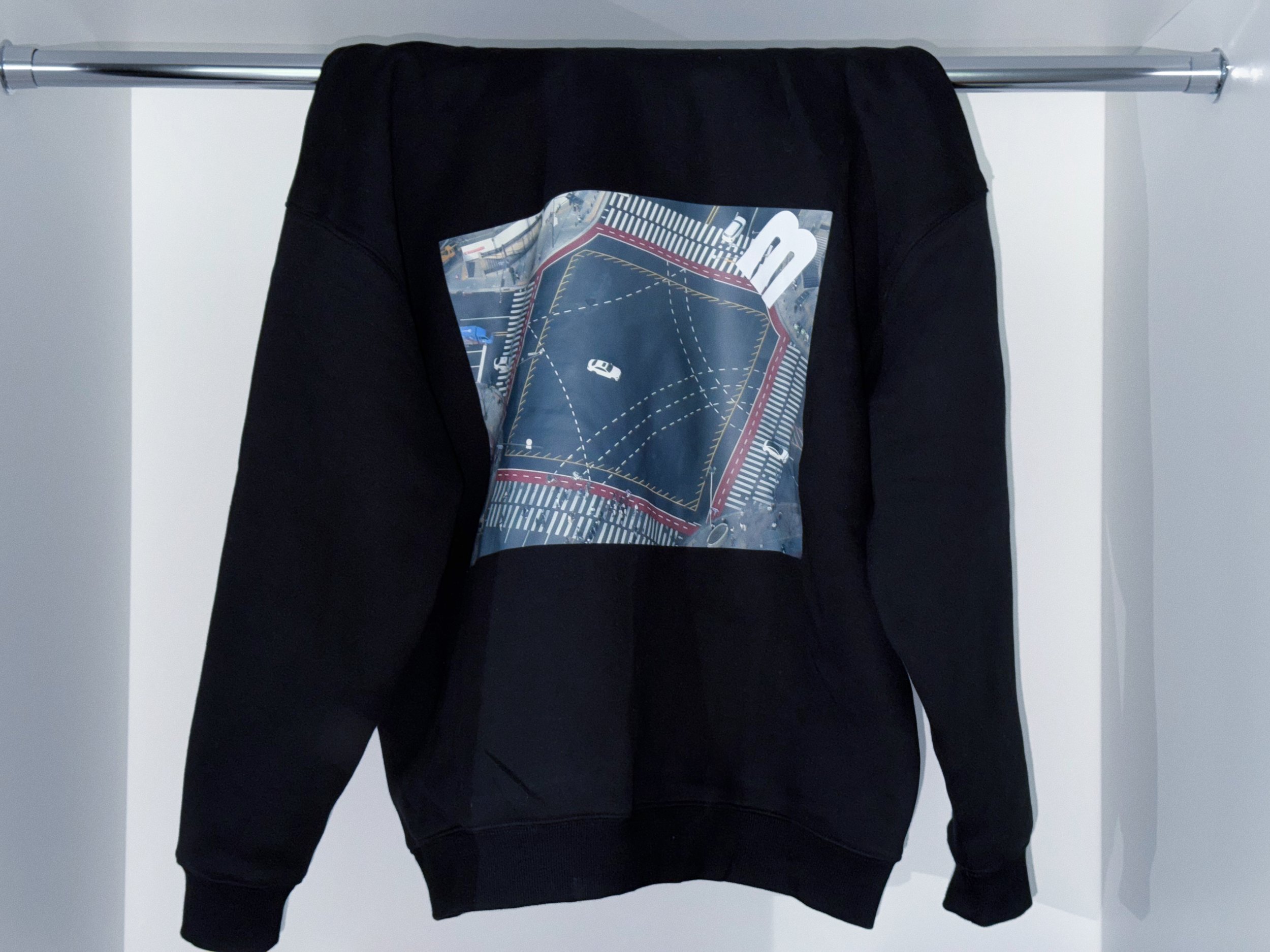 Black hoodie with a graphic print of a street intersection and cars, hanging on a gray metal rod against a plain light background.