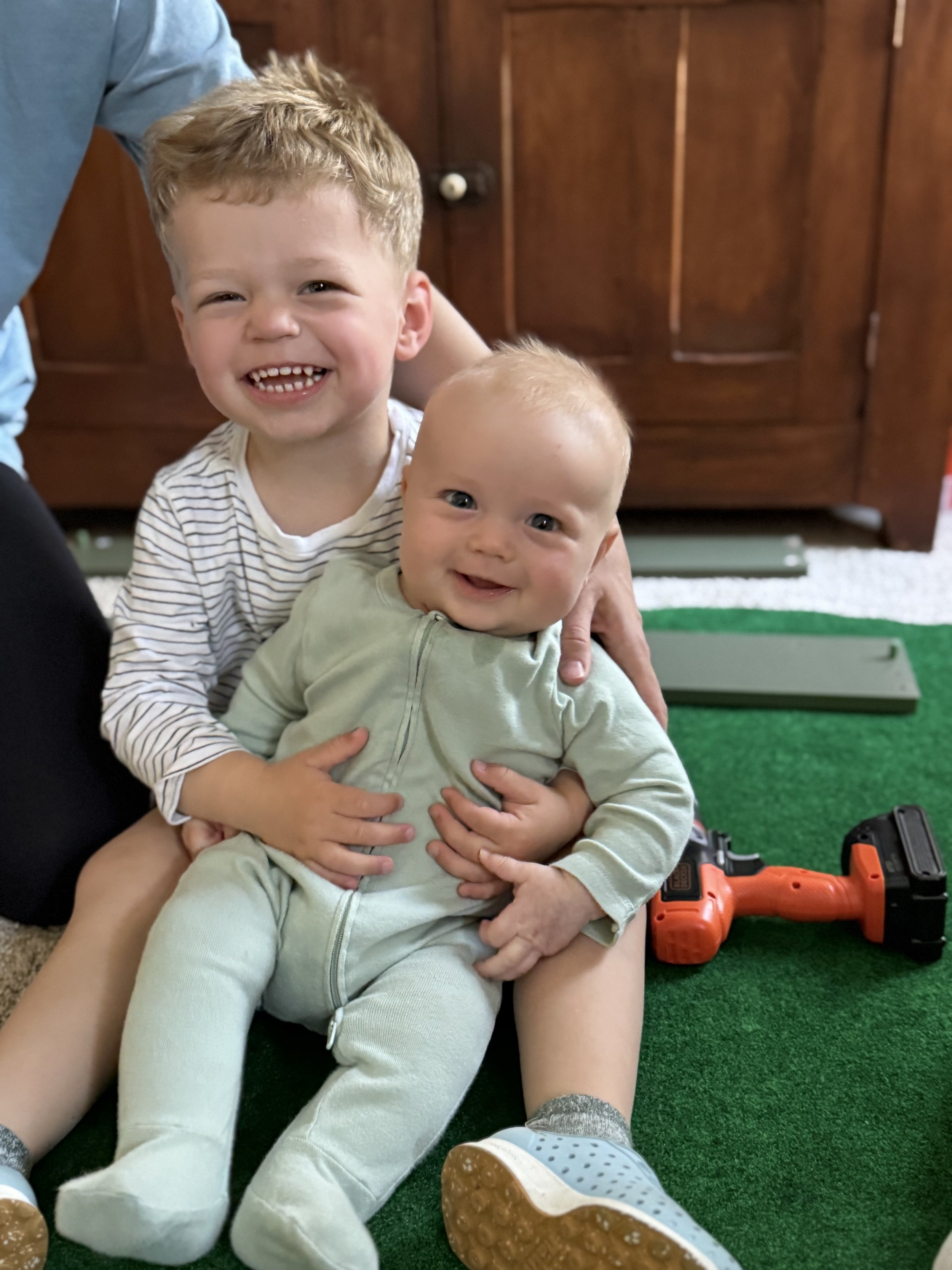Two young boys, one older with curly blonde hair and a striped shirt, and a younger with smooth blonde hair and a green outfit, sitting on a green carpet. The older boy is smiling broadly, showing his teeth, and the younger boy is also smiling. A toy tool is on the floor nearby, and a wooden cabinet is in the background.