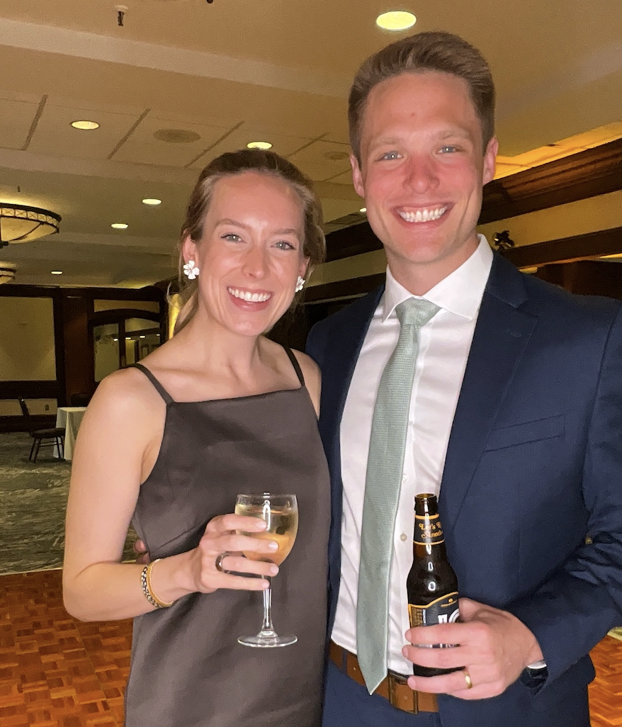A smiling woman in a dark sleeveless dress holding a glass of white wine, standing next to a smiling man in a dark blue suit with a light green tie holding a bottle of beer, at an indoor event or celebration.