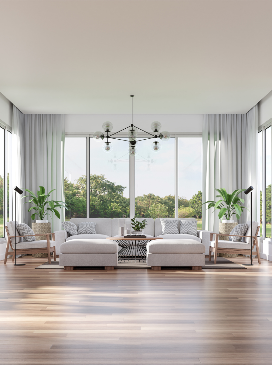 Bright living room with large windows, white curtains, white sectional sofa, two armchairs with pillows, potted plants, wooden floor, modern chandelier, and outdoor greenery visible through the windows.