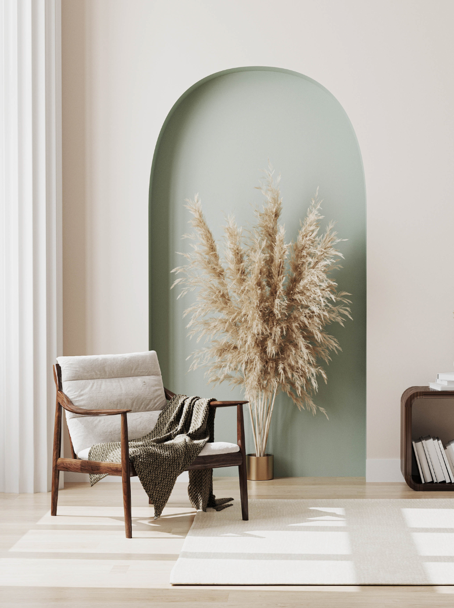 Modern living room corner with a white armchair, a blanket, a tall pampas grass arrangement in a vase, and a side table with books, against a partial curved green wall and light-colored curtains.