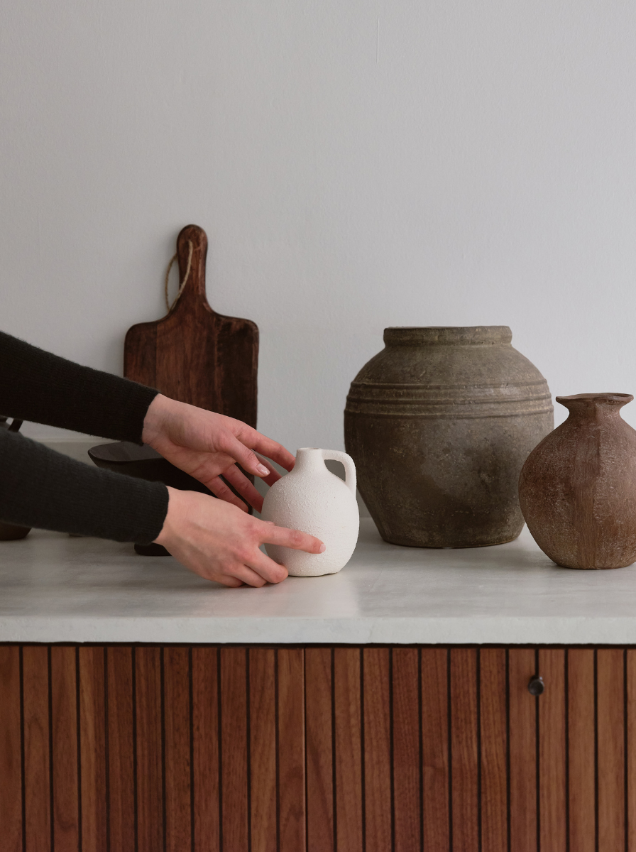 Person arranging small white ceramic pitcher on a wooden table with large antique clay and terracotta vases in the background.