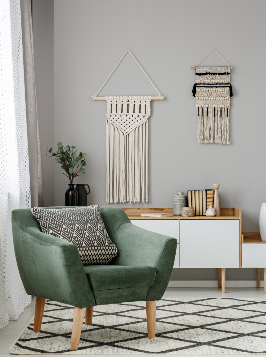 A cozy living room corner with a green armchair, patterned pillow, a black vase with greenery, a light wood and white cabinet holding books and decorative items, two neutral-toned wall hangings, and a window with sheer curtains.