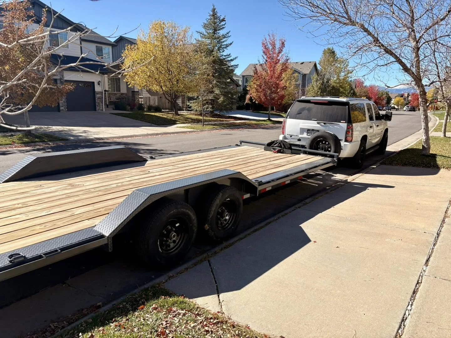 A white pickup truck attached to a flatbed trailer with wooden planks parked on a suburban street.
