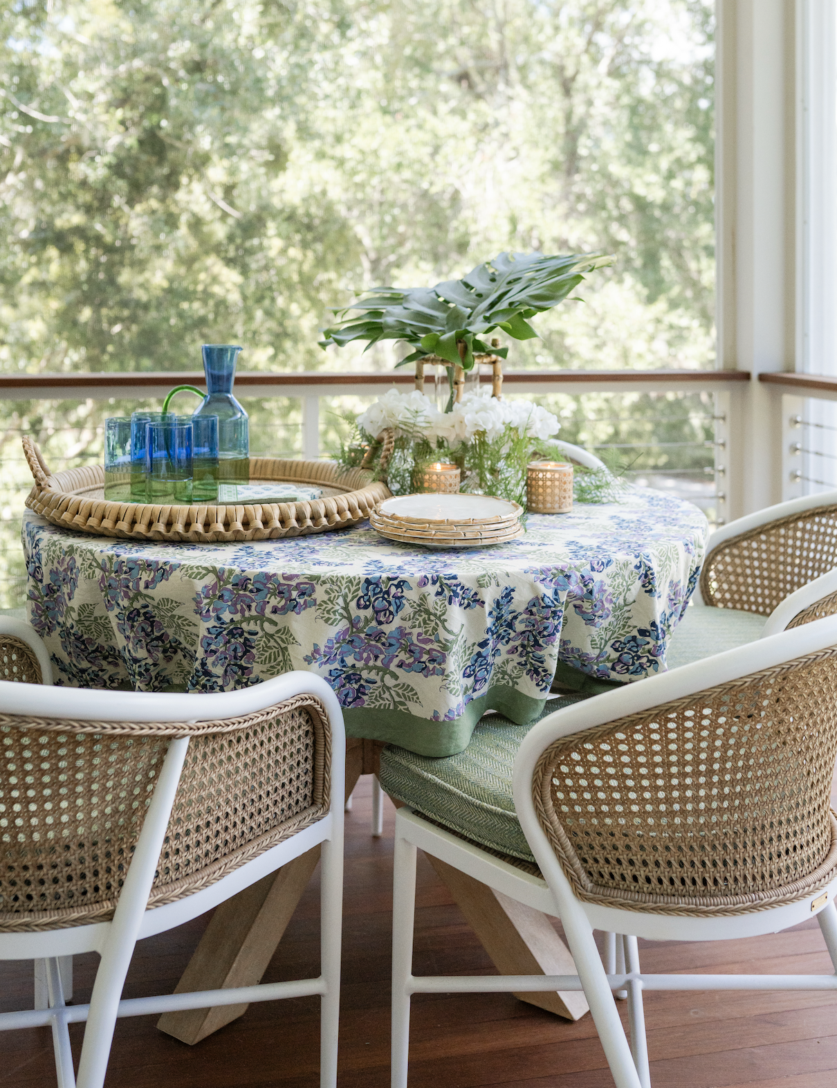 A round dining table with a floral tablecloth, decorated for a gathering, with wicker chairs around it. The table holds a large wicker tray with teal glasses and a blue vase, a stack of plates, and a centerpiece with green leaves, white flowers, two small candles, and natural light from large windows showing greenery outside.