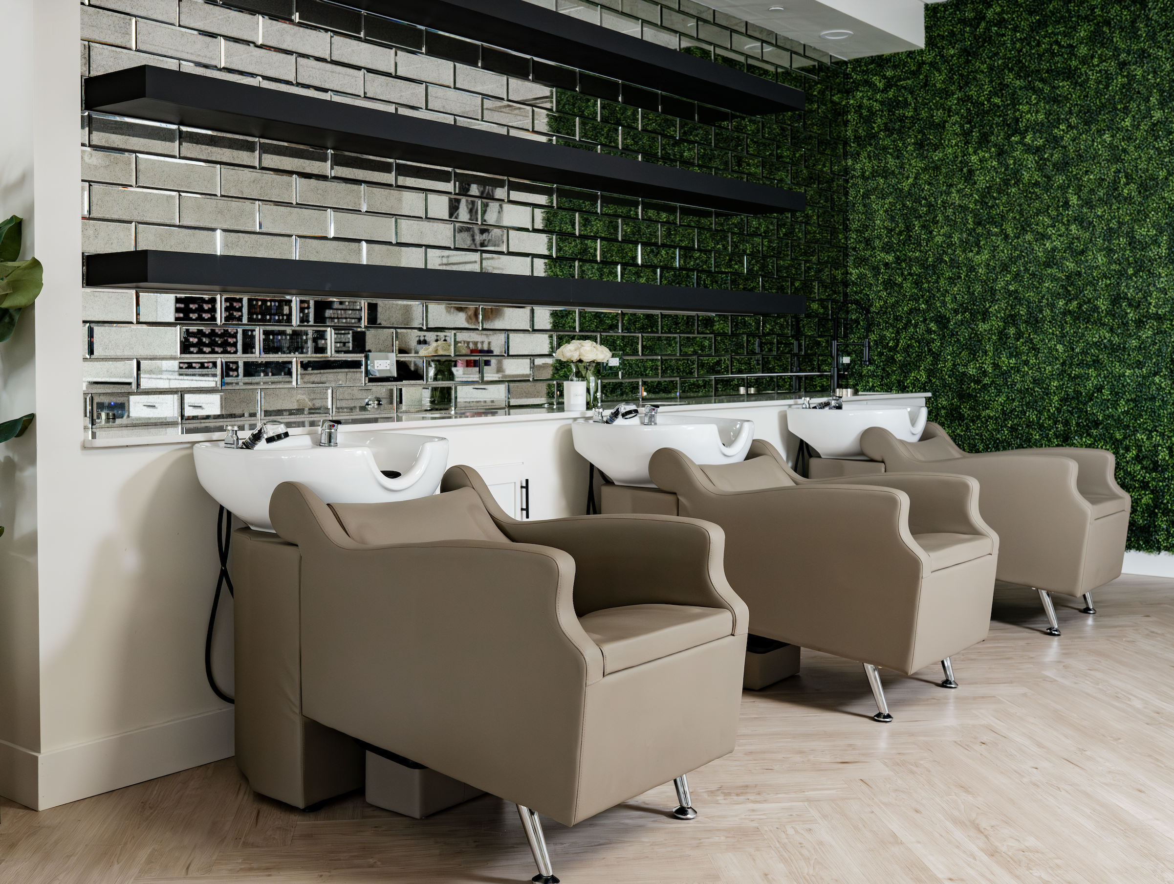 Hair salon with white washing basins and beige salon chairs in front of a green leafy wall, with a mirror and shelves behind.