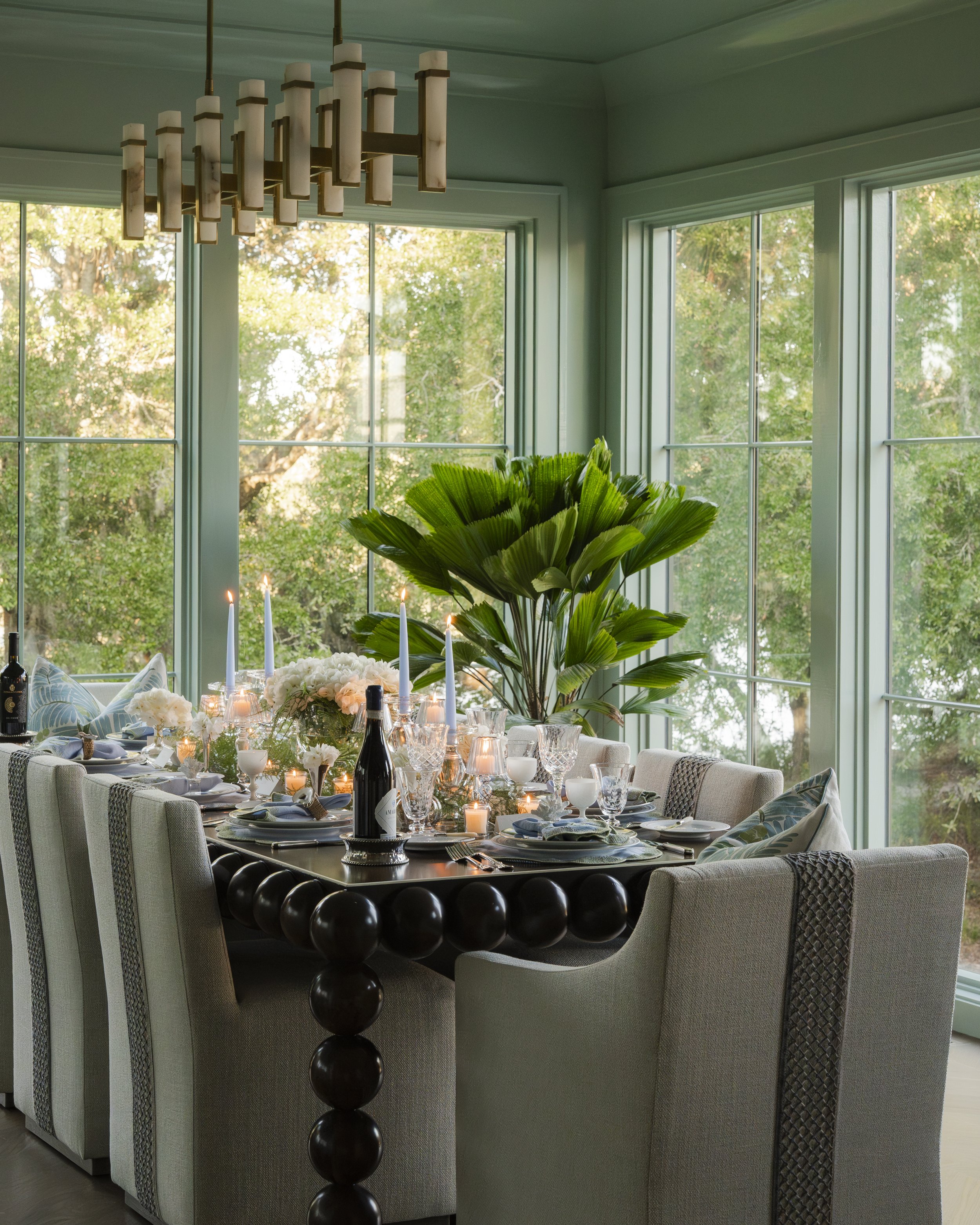 Interior of a sunlit dining room with large windows showing trees outside, decorated with a table set for a meal, candles, and a large green leafy plant.