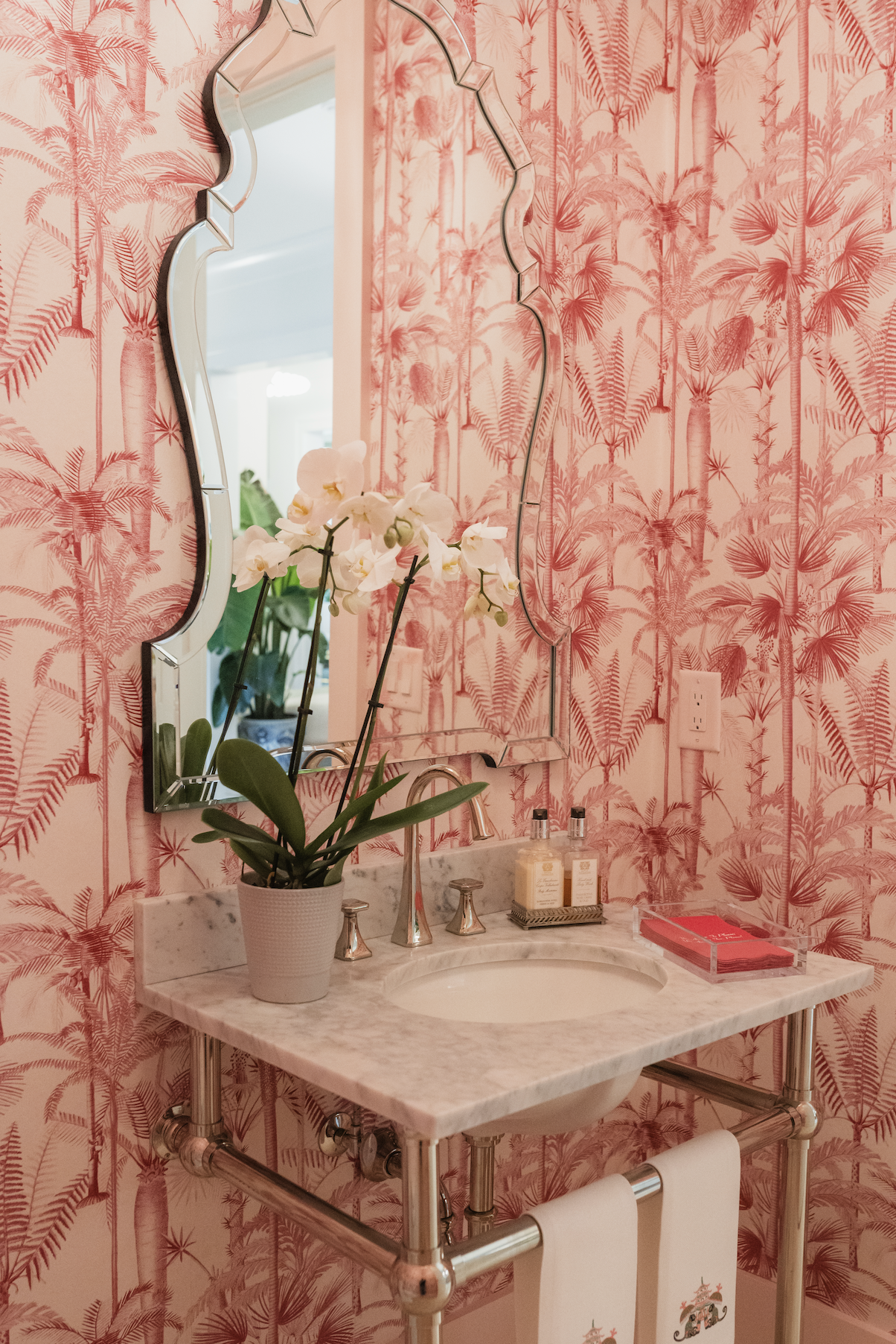 A bathroom with pink wallpaper featuring palm tree and tropical leaf patterns, a marble countertop with a built-in sink, a tall mirror with a decorative frame, a potted white orchid, soap and lotion bottles, and pink towels with holiday designs hanging below the counter.