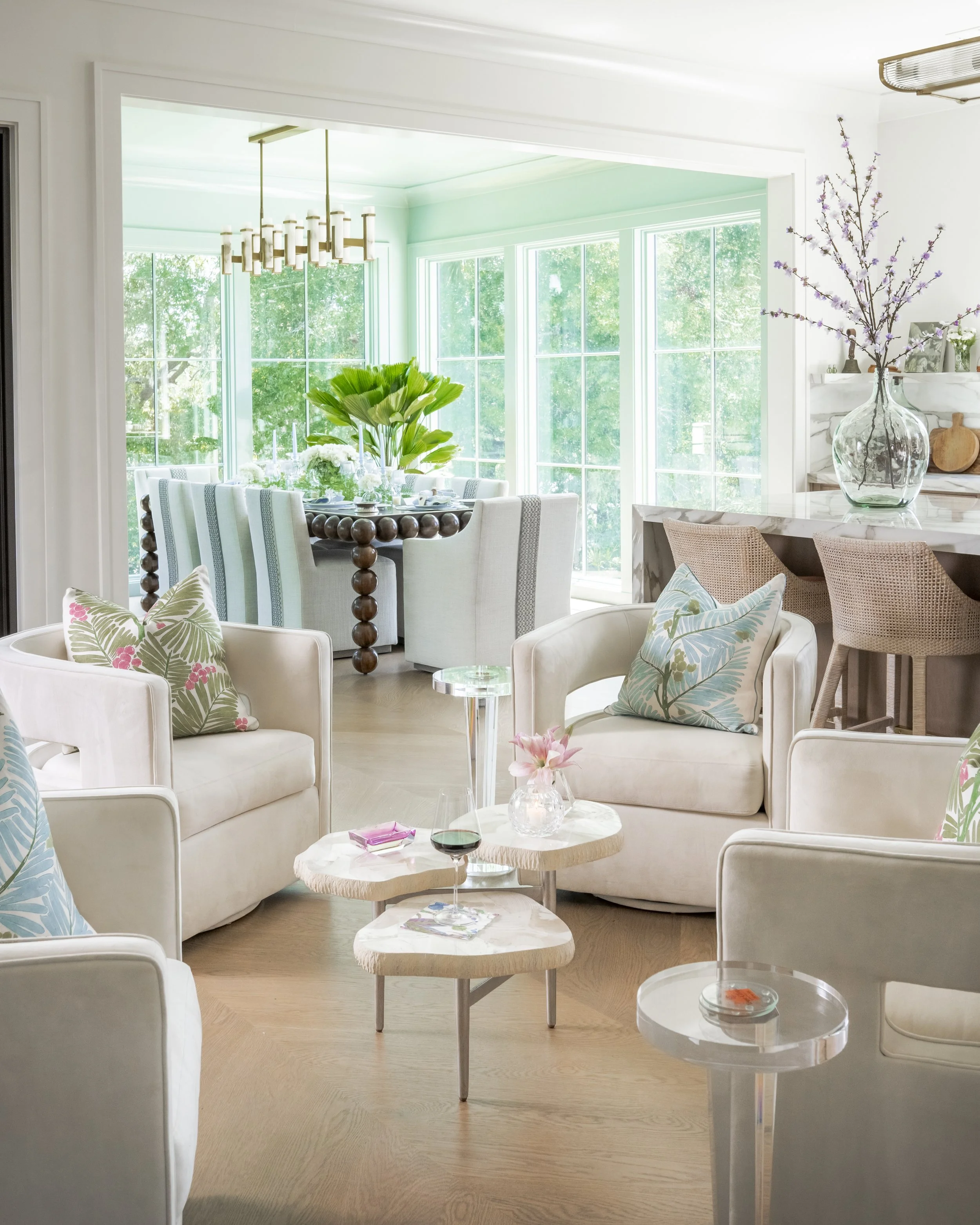 A bright living room with large windows and a view of green trees outside. The room has a dining table with a centerpiece, a chandelier, some armchairs with pillows, and small side tables with drinks and a flower vase.