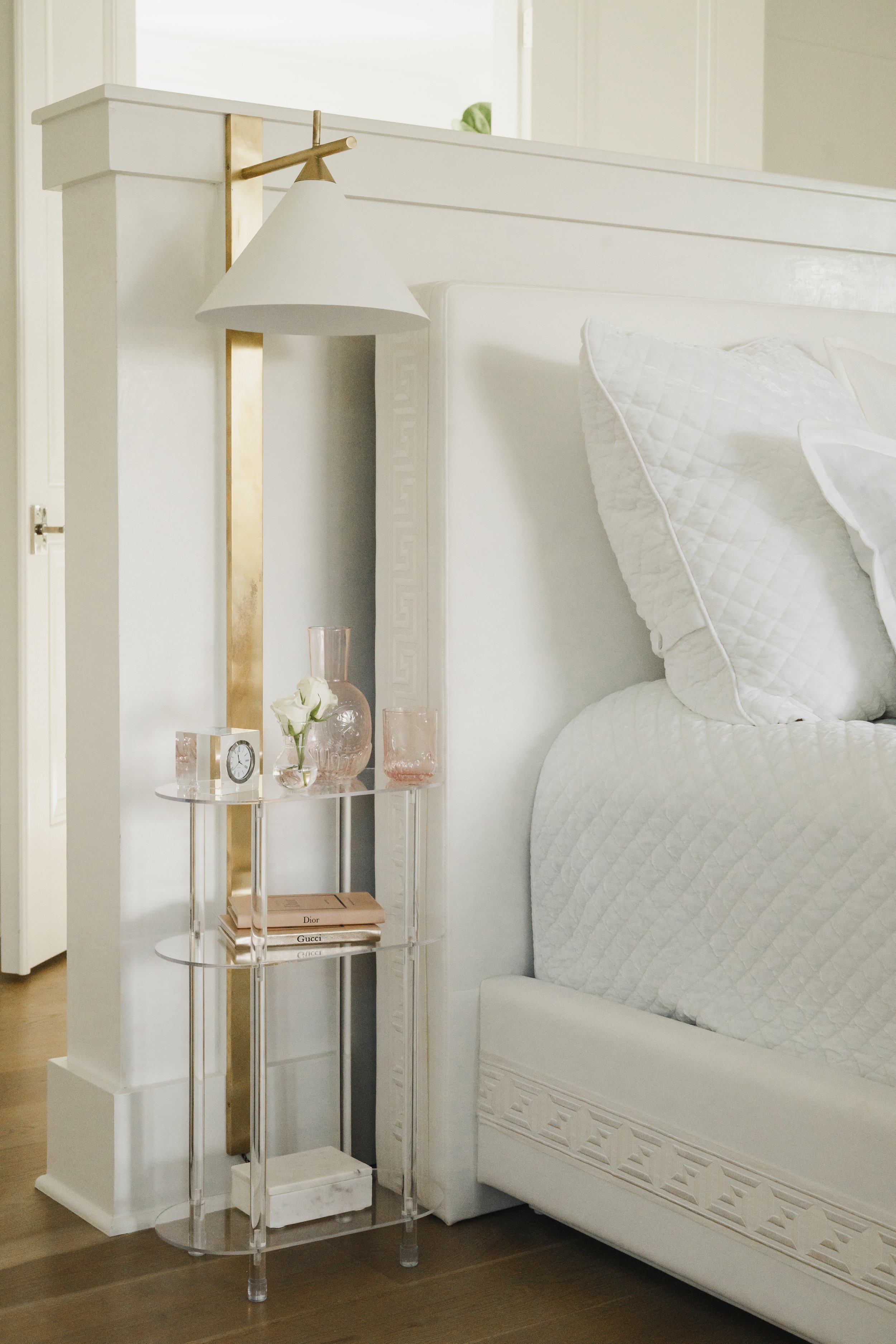 White bed with quilted pillows next to a transparent side table holding pink glasses, a clock, a pink vase with white flowers, and a stack of books, with a decorative wall lamp attached to a nearby white wall.