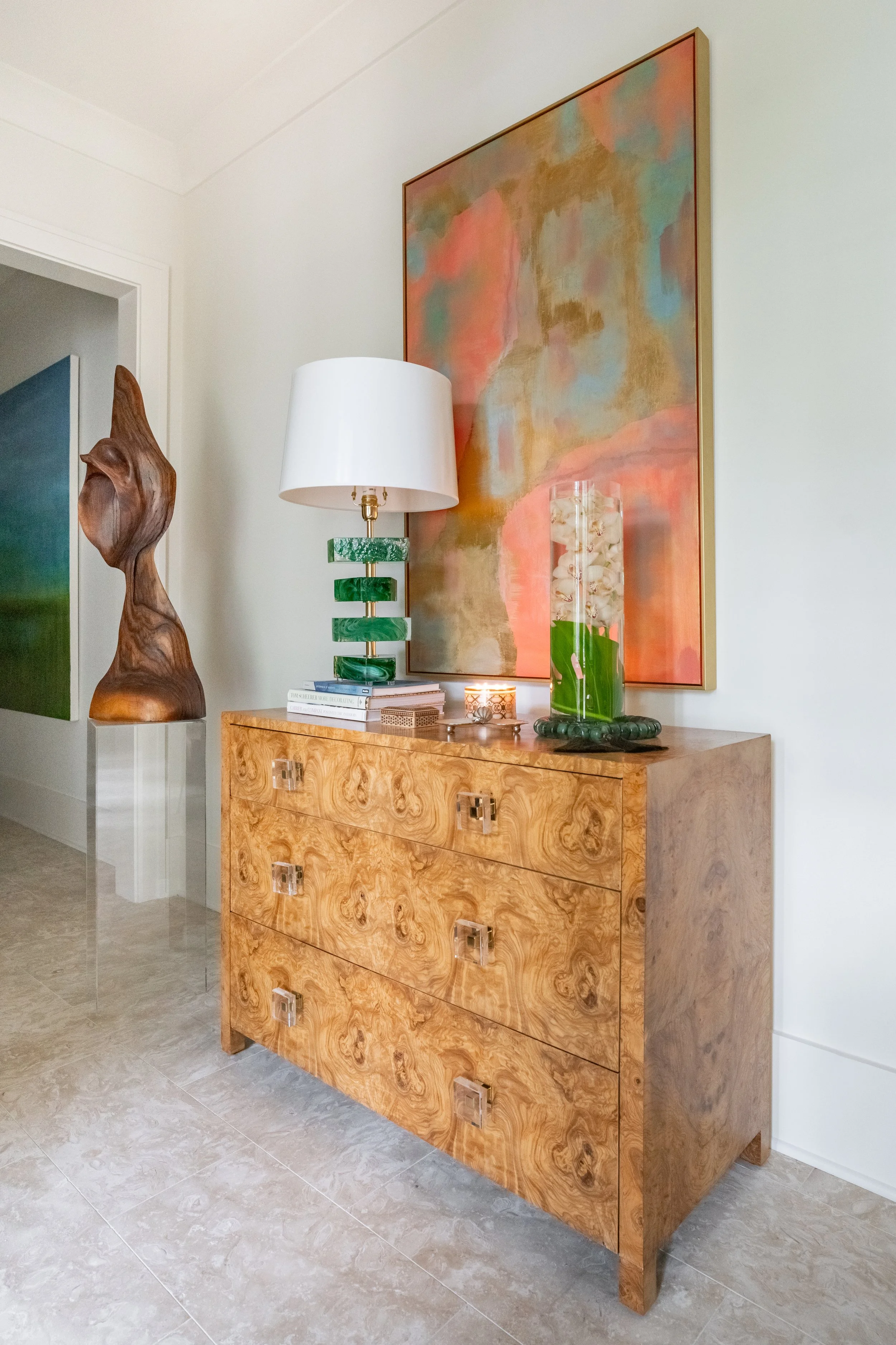 A wooden dresser with four drawers and square glass handles, decorated with a white lamp with a green marble base, a glass vase filled with white orchids, books, and a small lit candle. Behind, a large abstract painting with peach, blue, and gold tones hanging on a white wall.