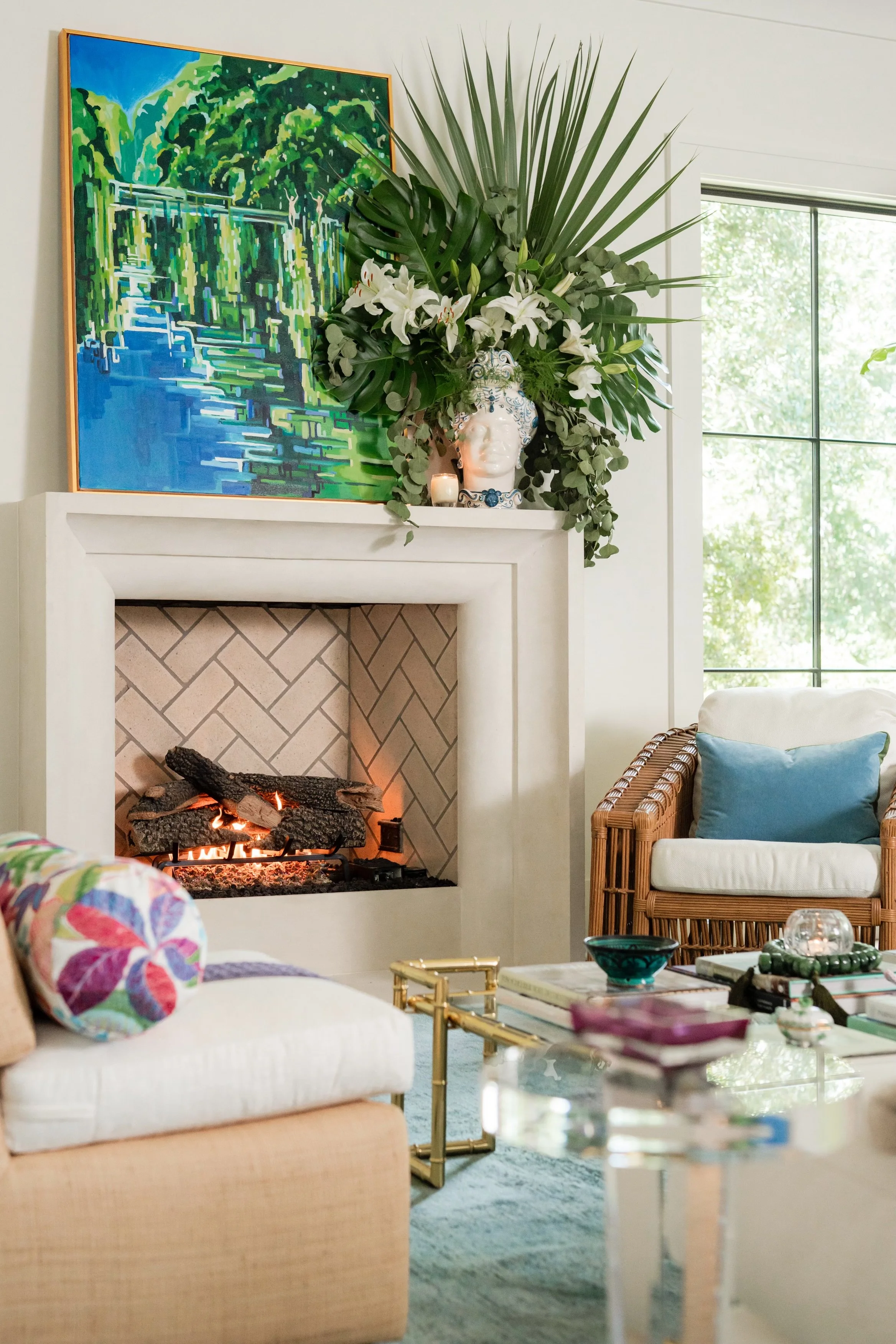 A living room with a fireplace, a colorful abstract painting of a water scene, a large arrangement of green tropical plants and white lilies on the mantelpiece, and a cozy armchair with a blue pillow near a window with mountain views.