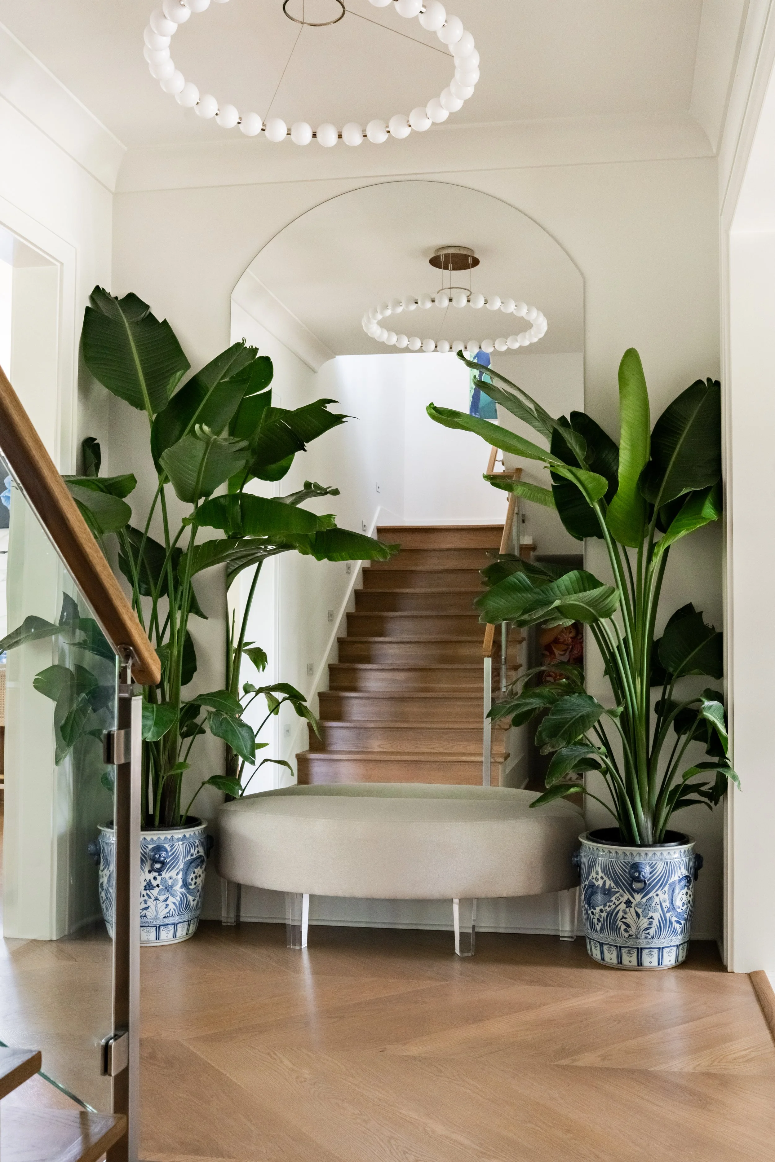 A hallway with wooden stairs and two large potted plants in blue and white ceramic pots on either side of a round, light-colored bench. The stairs lead to an upper floor, with two circular chandeliers hanging from the ceiling.