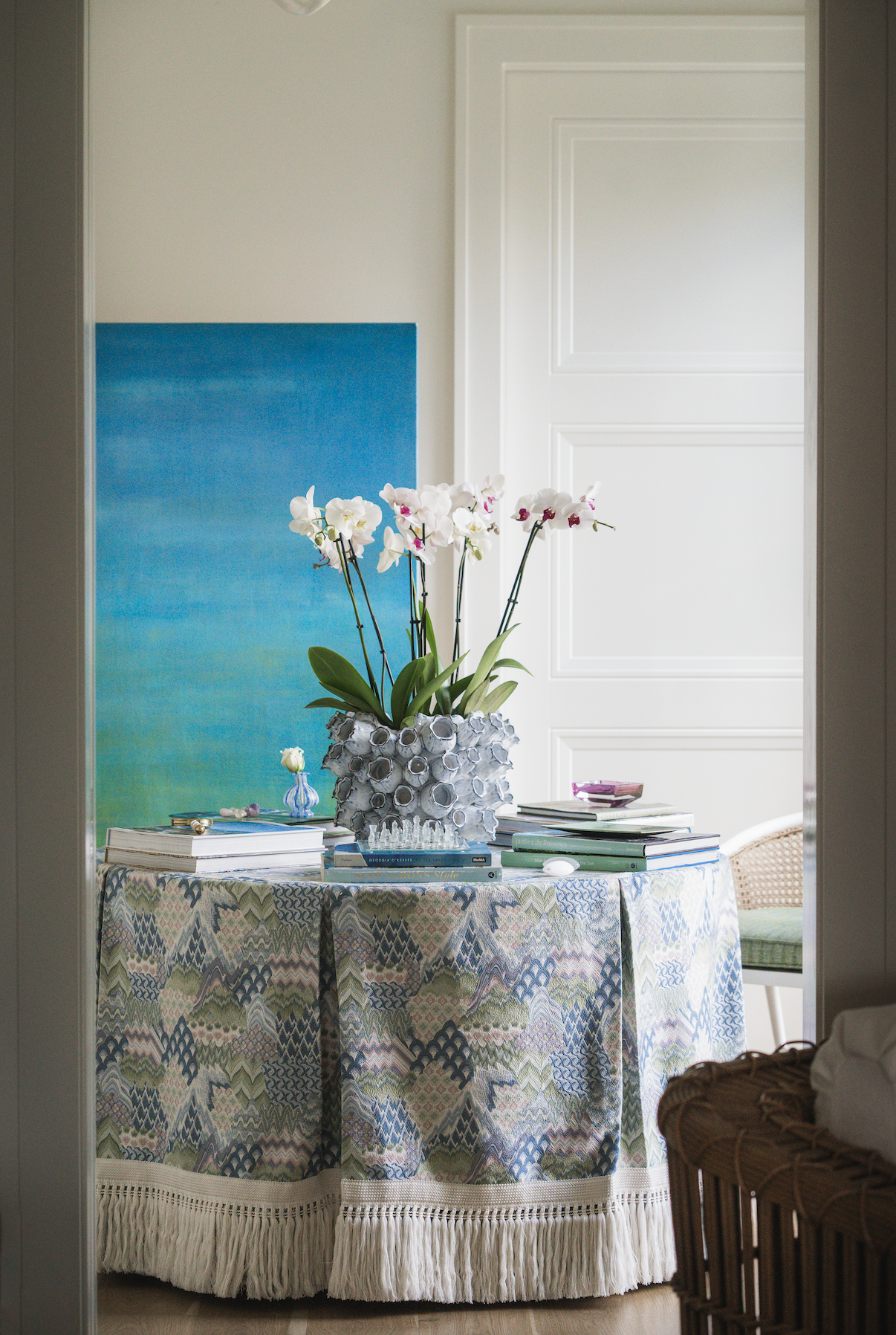 A round table covered with a colorful, patterned tablecloth with fringe, topped with a large white vase of orchids, books, and decorative objects. A blue abstract painting is on the wall behind. The scene is viewed through a doorway into a bright room with a white door and chair.