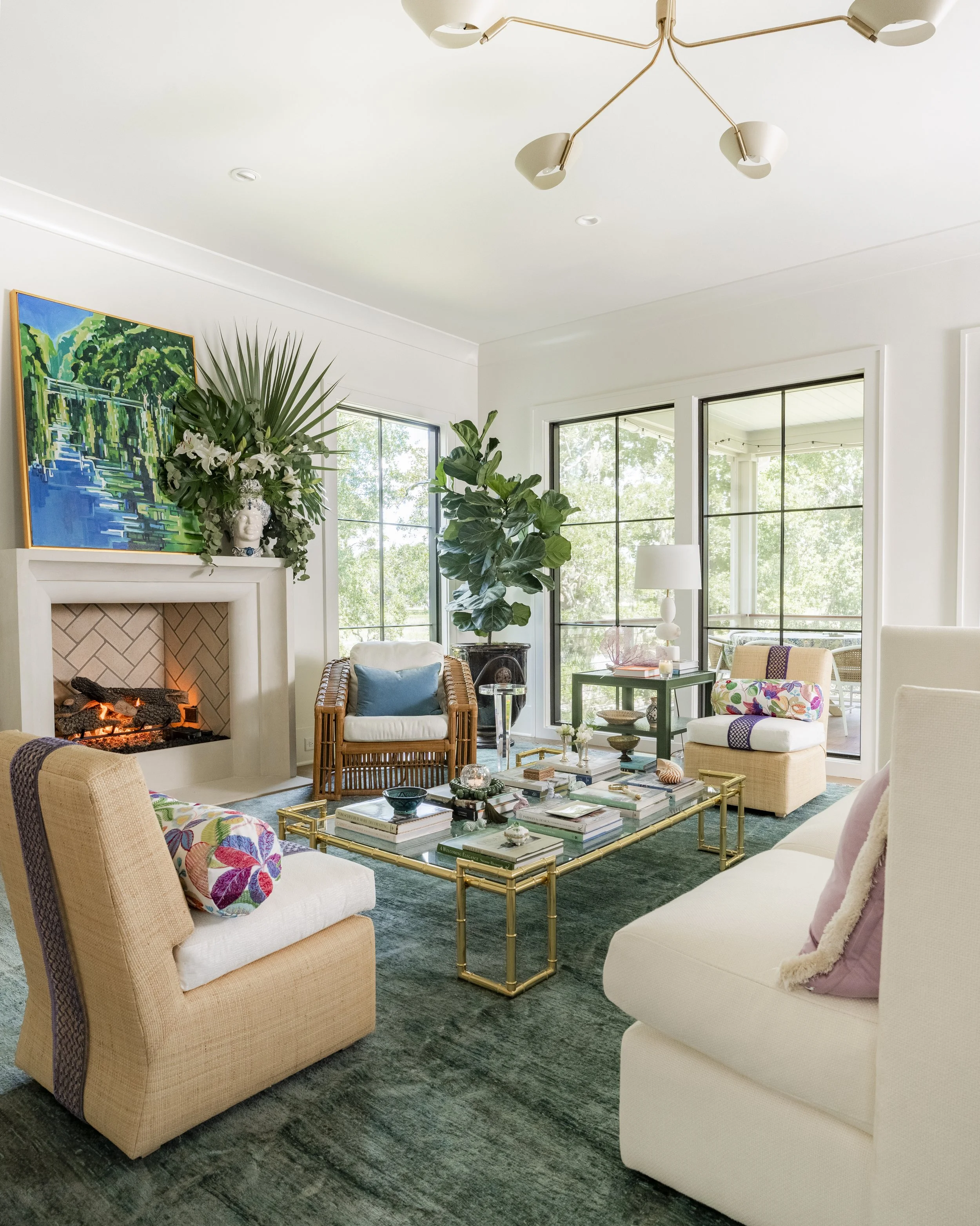 A bright living room with large windows, a fireplace, and modern chairs. Decor includes a gold coffee table, colorful cushions, a large green plant, and artwork above the fireplace. There's a ceiling light fixture.