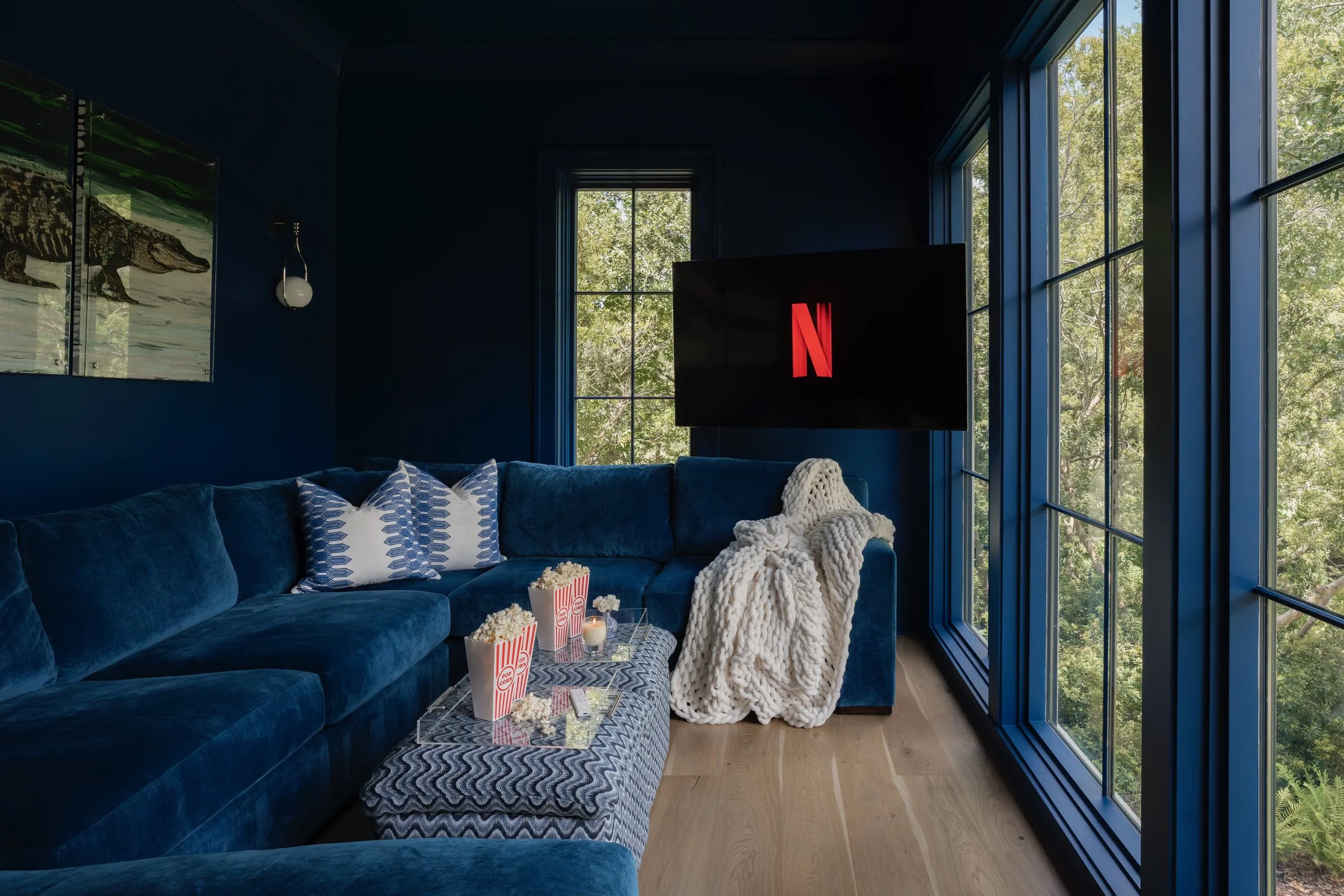 Living room with plush blue velvet sofa, decorative pillows, and a coffee table with popcorn, popcorn buckets, and candles. Large windows reveal lush green trees outside. A black TV screen with a red Netflix logo is mounted on the wall. Art prints featuring a crocodile and nature scene hang on the dark blue wall, and a modern spherical light fixture is on the wall.