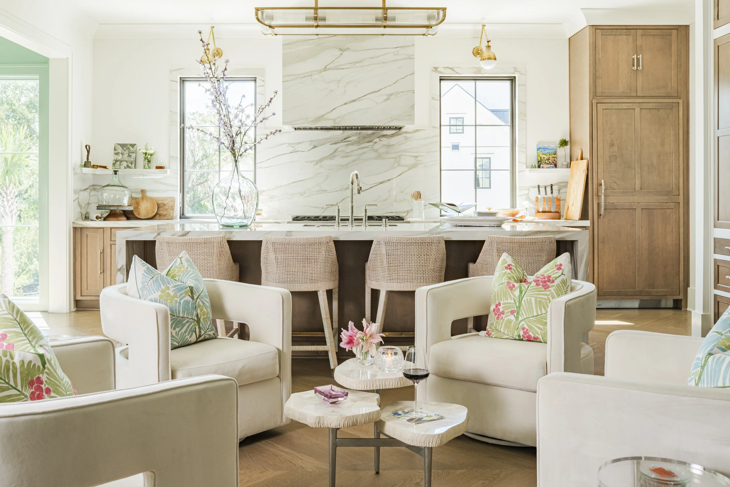 A bright, modern kitchen with white marble backsplash and countertops, natural wood cabinets, large windows overlooking greenery, and a dining area with white chairs and floral cushions. Decor includes a large glass vase with branches, books, plants, and decorative items.
