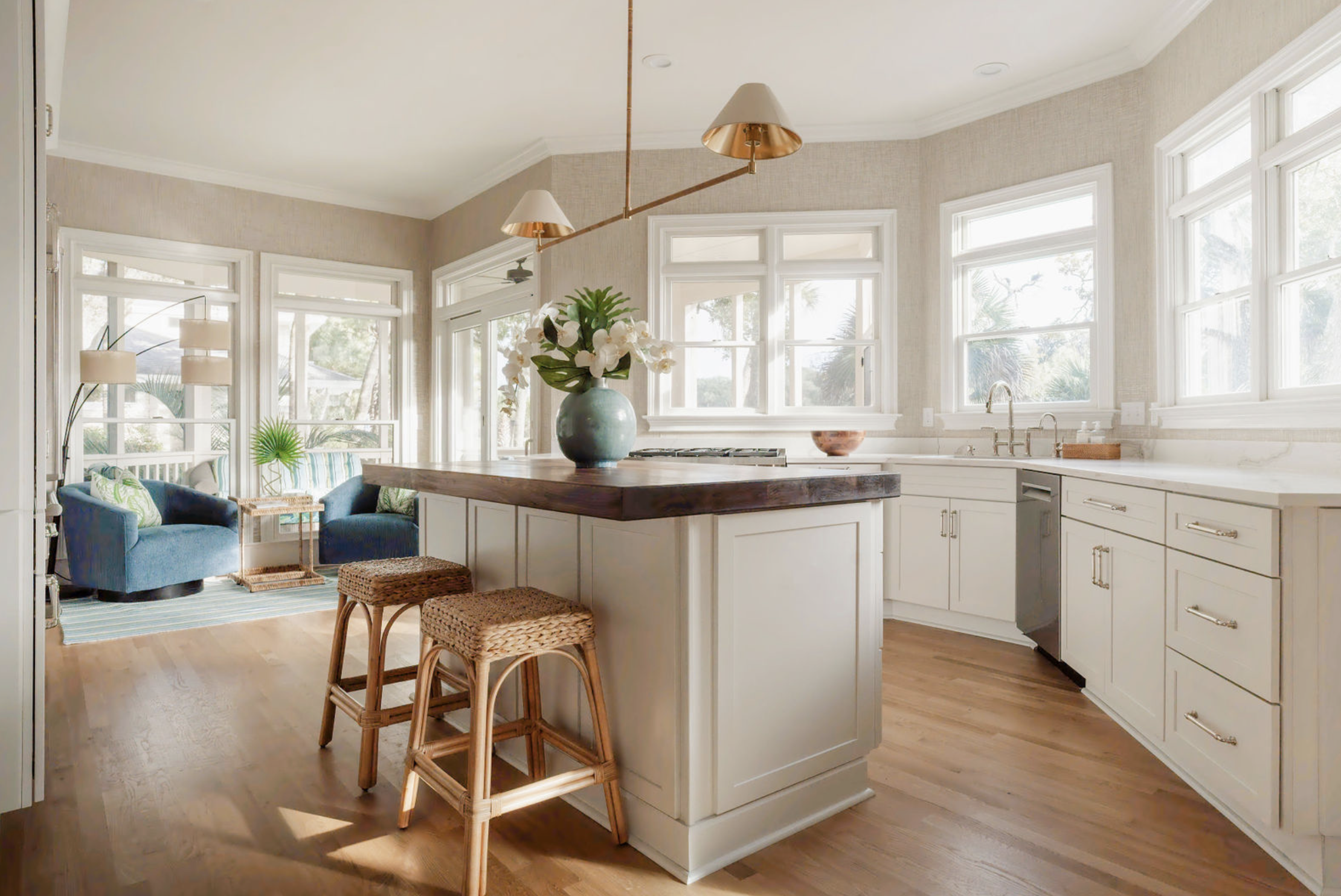 Sunlit kitchen with white cabinetry, a large island with a dark wooden countertop, and a vase of white flowers. In the background, a cozy seating area with blue armchairs and a striped rug is visible.