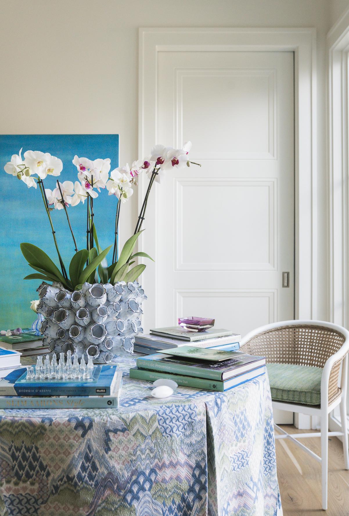 A dining area with a table covered by a colorful patterned tablecloth, decorated with a white textured ceramic vase holding white orchids with pink accents, and stacks of books. There is a white chair with a woven cane backrest and a cushioned seat. In the background, a closed white door and a window are visible.