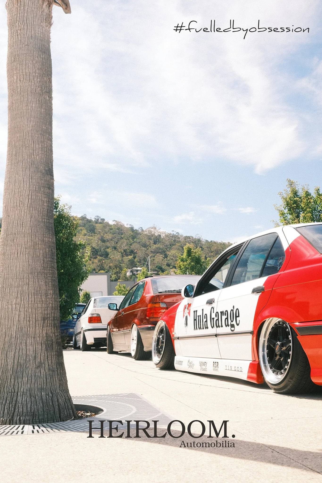 Car parked in a parking lot with a large tree in the foreground, behind which there are other vehicles and a hillside covered with trees under a partly cloudy sky. There is text on the image that says 'HEIRLOOM Automobilia' and some hashtags in the sky area.