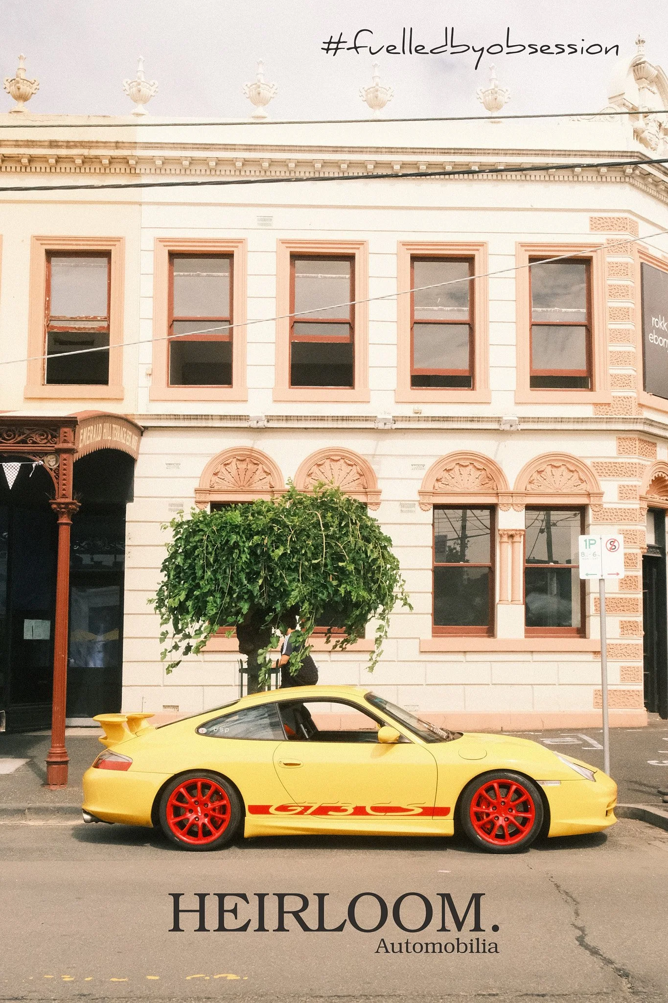 A yellow Porsche sports car with red wheels and a racing stripe parked on a city street in front of a beige building with multiple windows. There is a green tree in front of the building and a red street lamp on the left side. The image has text overlay that says '#fueledbyobsession' at the top and 'HEIRLOOM. Automobilia' at the bottom.