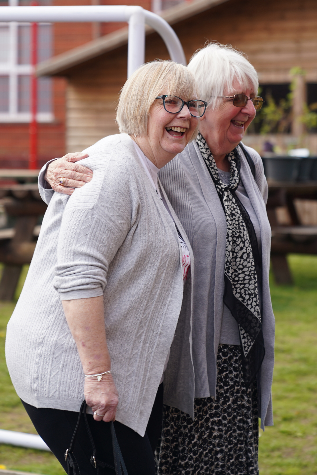 Two elderly women smiling and standing close together outdoors, one with light brown hair and glasses, the other with white hair and sunglasses, with a wooden building in the background.