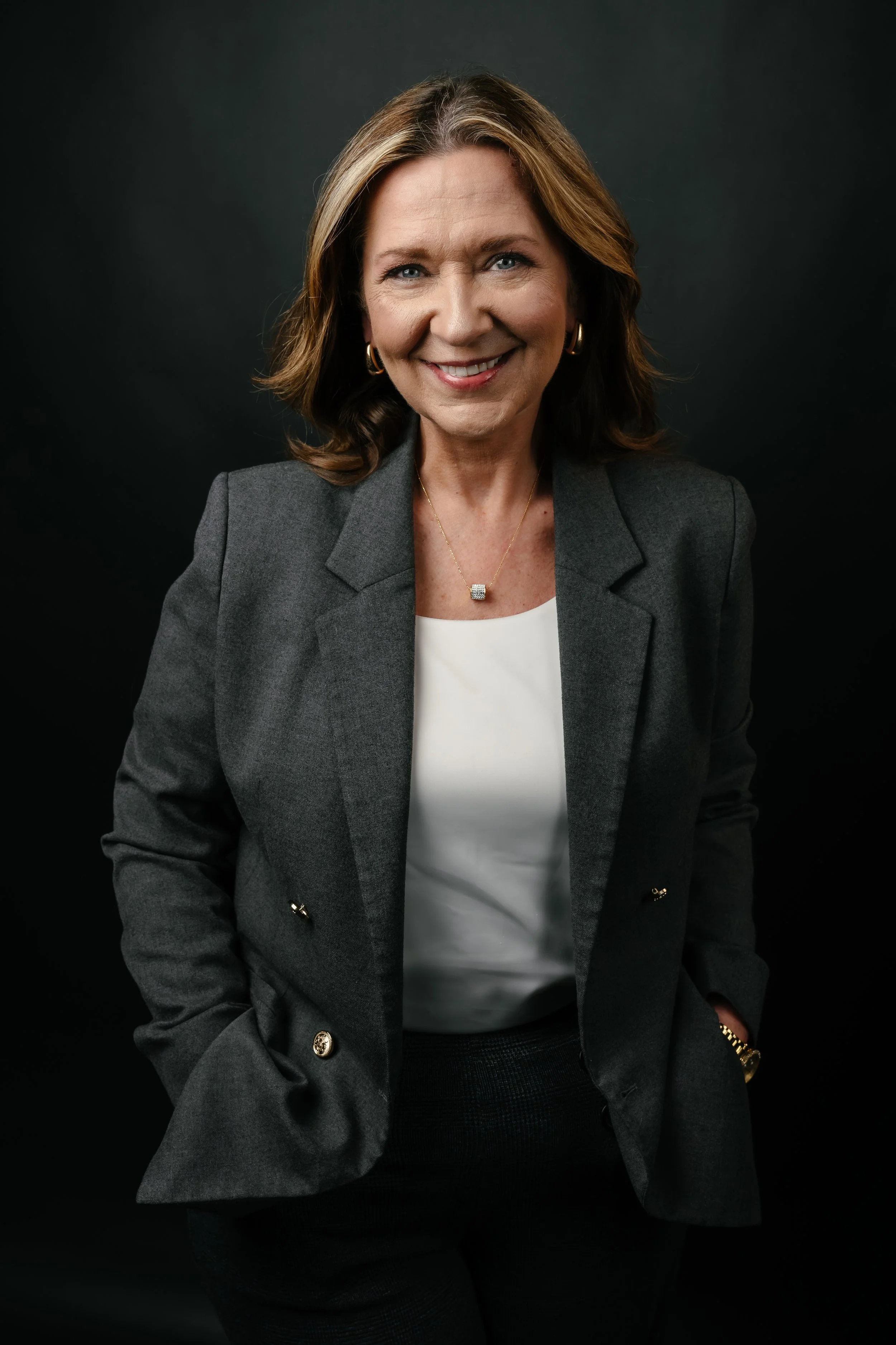 Smiling woman with light brown hair in a professional gray blazer and white top, posing against a dark background.