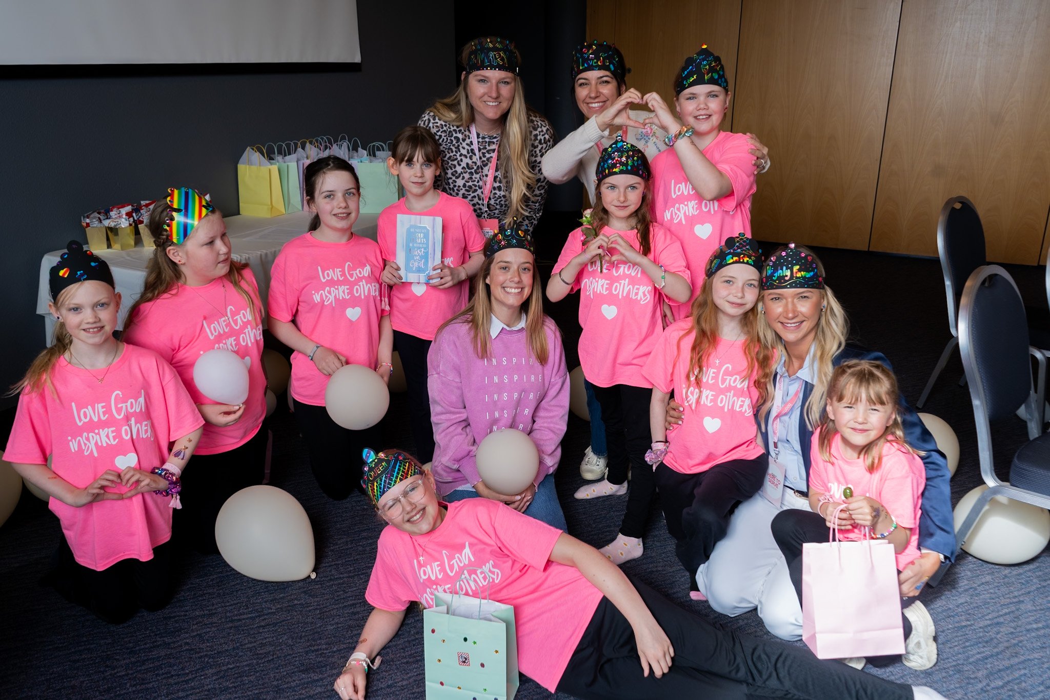 Group of young girls and women at a celebration, many wearing pink t-shirts with the phrase 'Love God, Inspire Others', some wearing colorful hats, and holding balloons and gift bags, in a room with dark walls and wooden doors.
