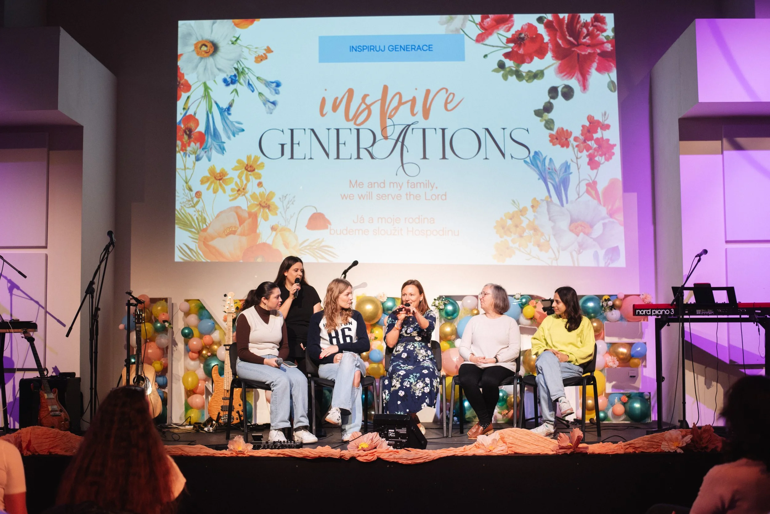 Women sitting on a stage decorated with balloons in front of a large screen, during a panel discussion or presentation. The large screen displays a floral-themed slide with the words 'Inspire Generations' and additional text in English and another language, with microphones, guitars, and musical equipment visible.