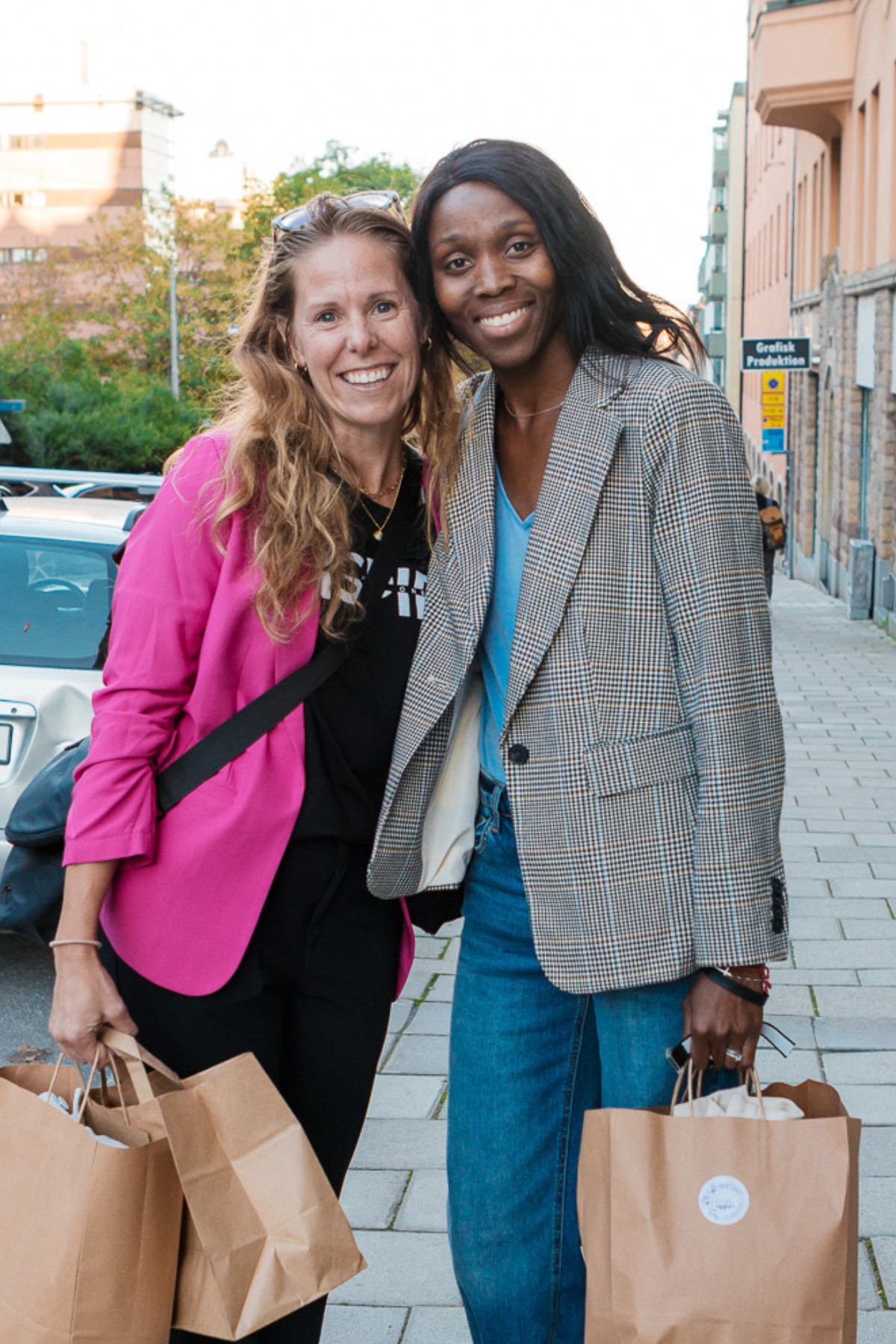Two women standing on a city sidewalk holding shopping bags and smiling at the camera. One woman has long red hair and is wearing a bright pink blazer, the other has long black hair and is wearing a checked blazer and blue jeans.