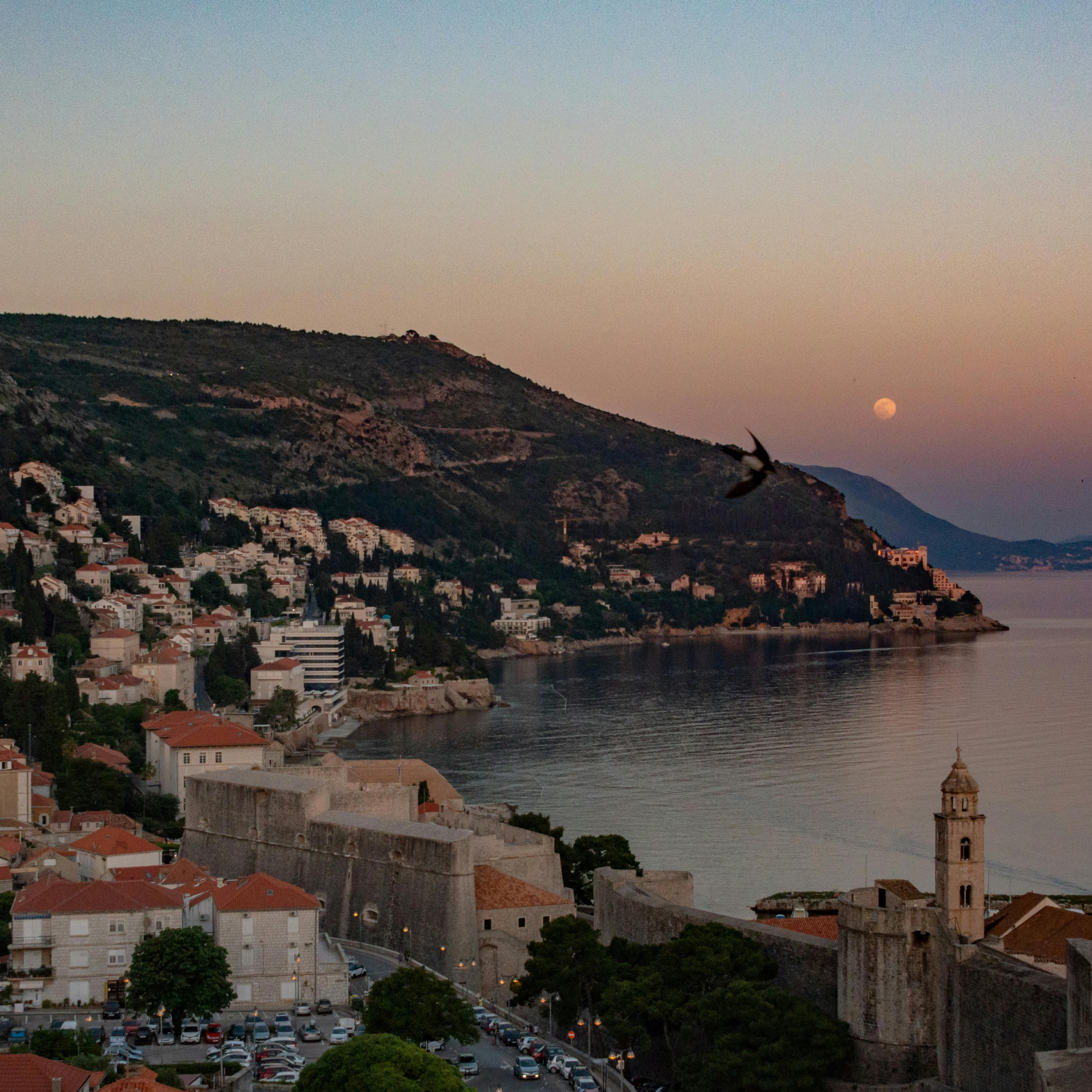 A coastal hillside town at dusk with white and red-roofed buildings, a stone fortress wall at the foreground, a church steeple, and the moon visible near the horizon over calm water.
