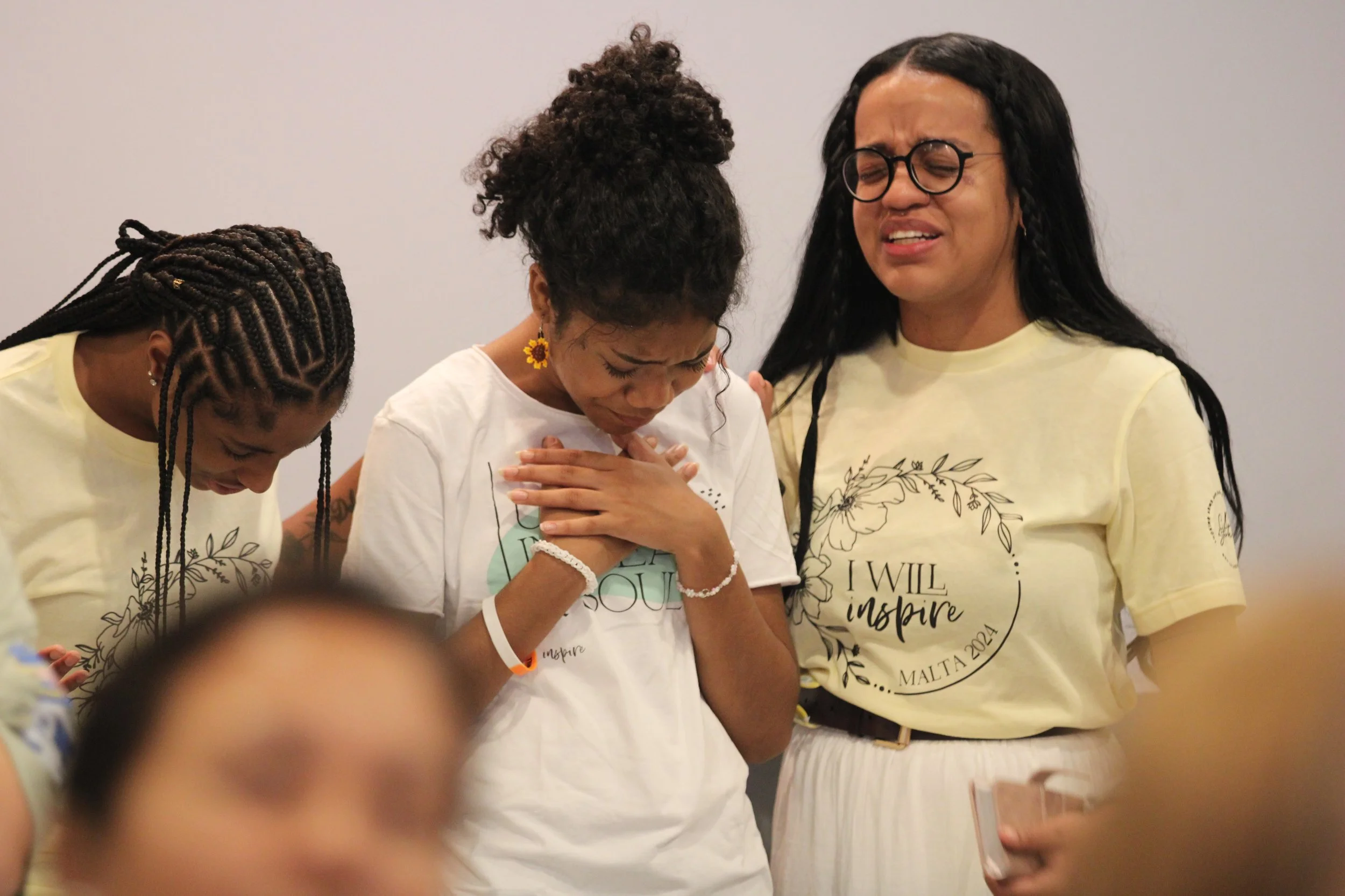 Three women with braids and wearing yellow T-shirts with inspirational messages are in a moment of prayer or reflection, with one woman holding her hands over her heart and closed eyes.