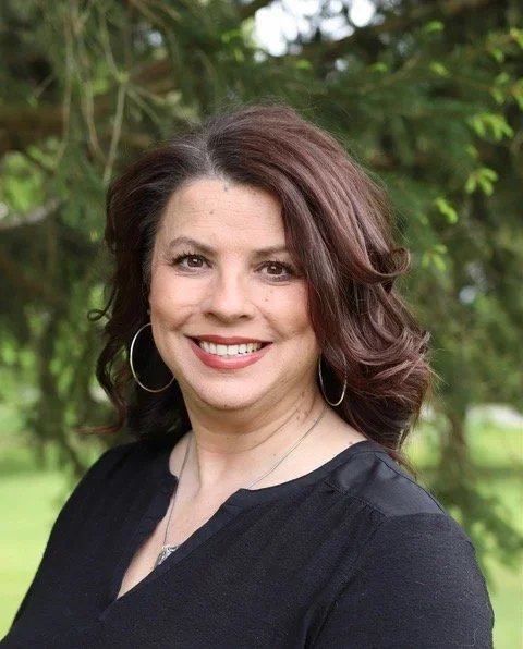 A woman with shoulder-length dark red hair, hoop earrings, and a black top, standing outdoors with greenery in the background.
