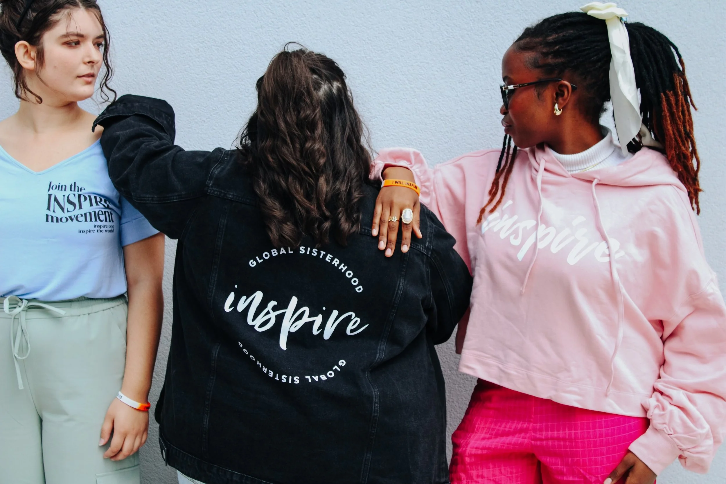 Four women standing against a plain wall. The women are outdoors and are wearing casual outfits with messages of inspiration on their clothes and accessories. They are touching, hugging, or standing close to each other, indicating camaraderie or friendship.