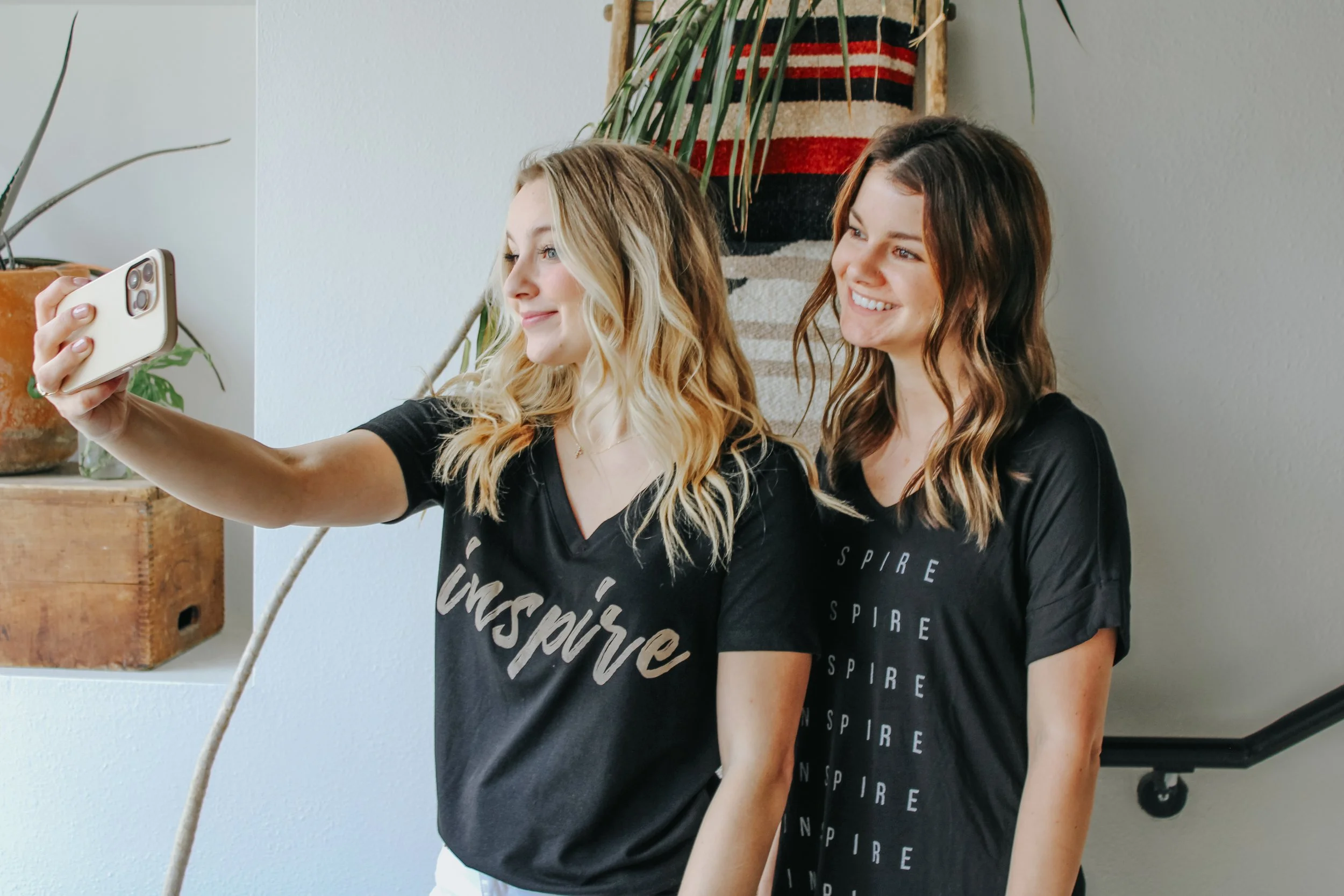 Two women taking a selfie together, standing indoors, smiling, with plants and wall art in the background.