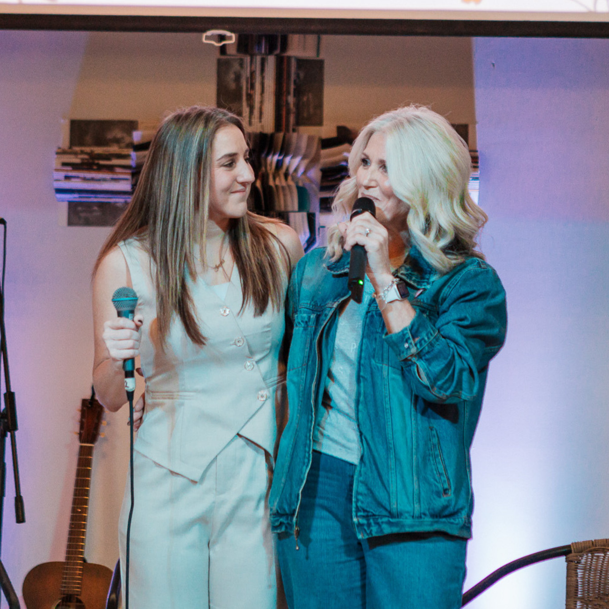Two women are standing close together, smiling and engaging in conversation. One woman is holding a microphone. The background shows a wall with framed artwork, and there are some chairs and a guitar visible in the setting.
