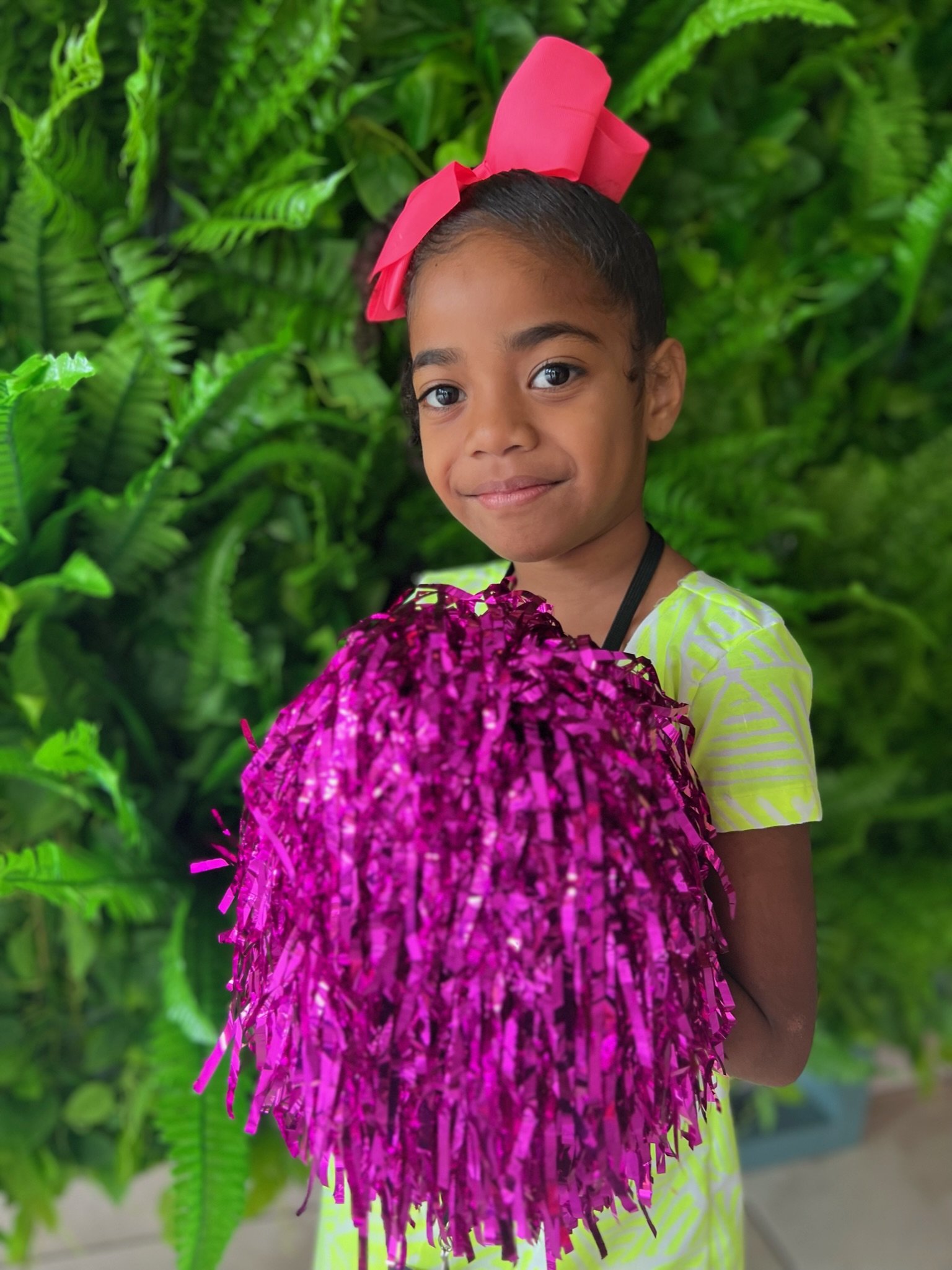A young girl with dark hair, wearing a pink bow headband, holding pink shiny pom-poms, standing in front of lush green foliage.