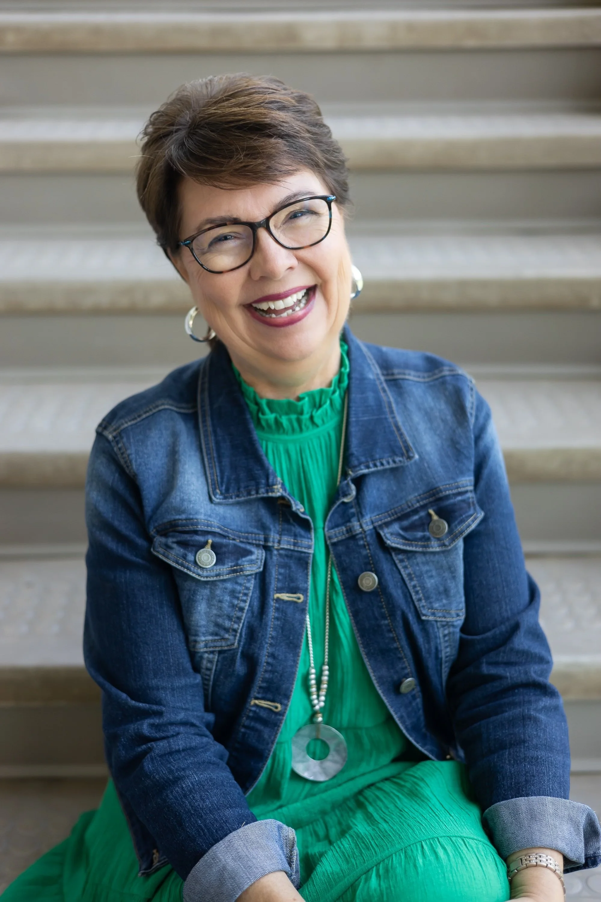 A smiling woman with short brown hair, glasses, and jewelry, sitting on stairs wearing a denim jacket and a green dress.