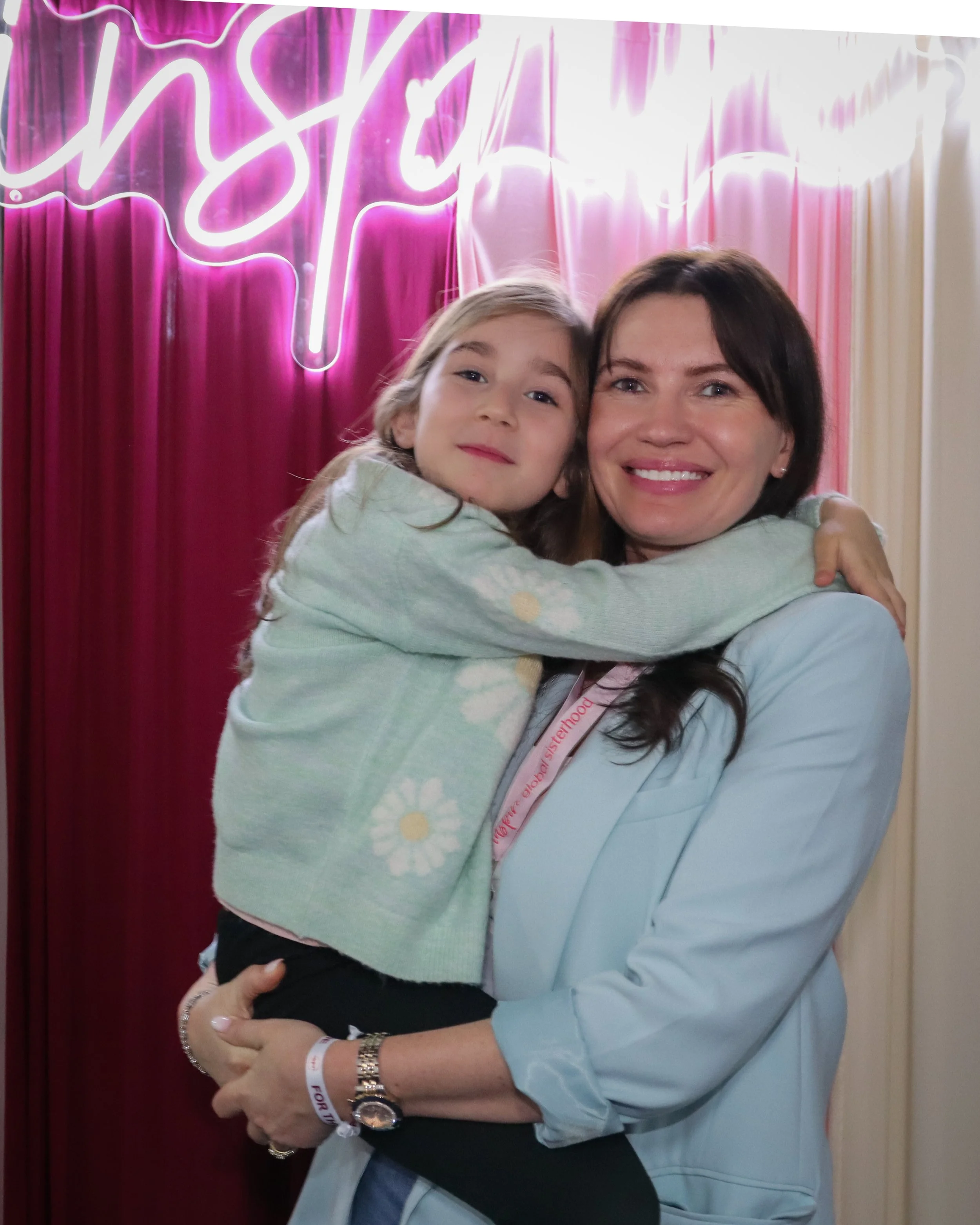 A woman and a young girl are hugging and smiling at the camera. The woman has dark hair and is wearing a light blue shirt. The girl has blonde hair and is wearing a hoodie with a flower pattern. Behind them is a pink velvet curtain and a white neon sign.