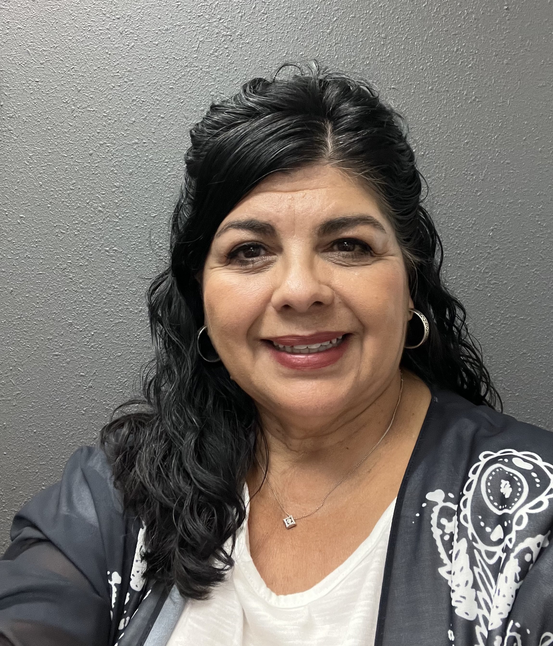 Close-up of a woman smiling with curly black hair, wearing earrings, a necklace, a white top, and a black jacket with a white paisley pattern, standing against a gray textured wall.