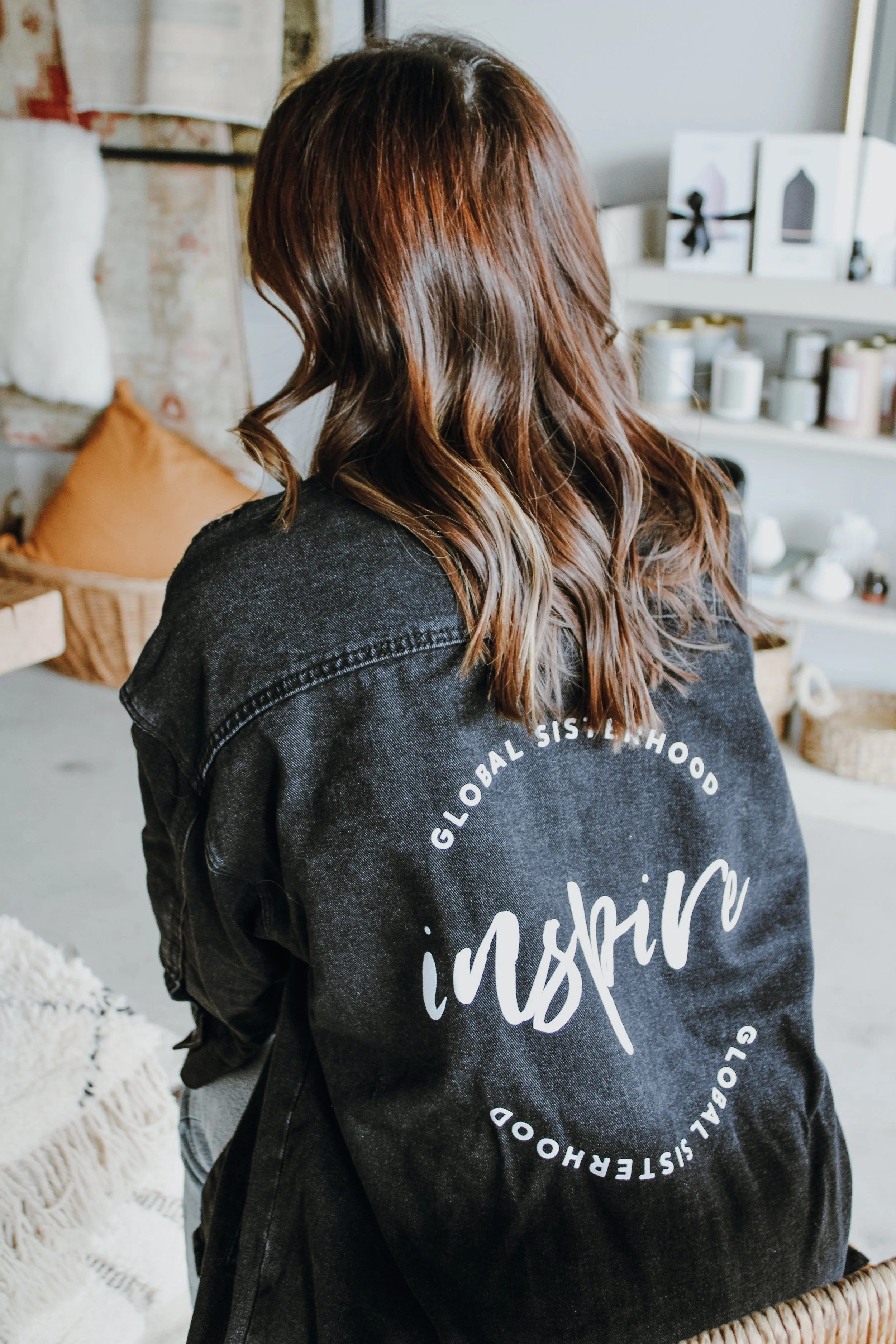 Back view of a woman with wavy, reddish-brown hair wearing a black denim jacket with white circular text, in a cozy, decorated room.