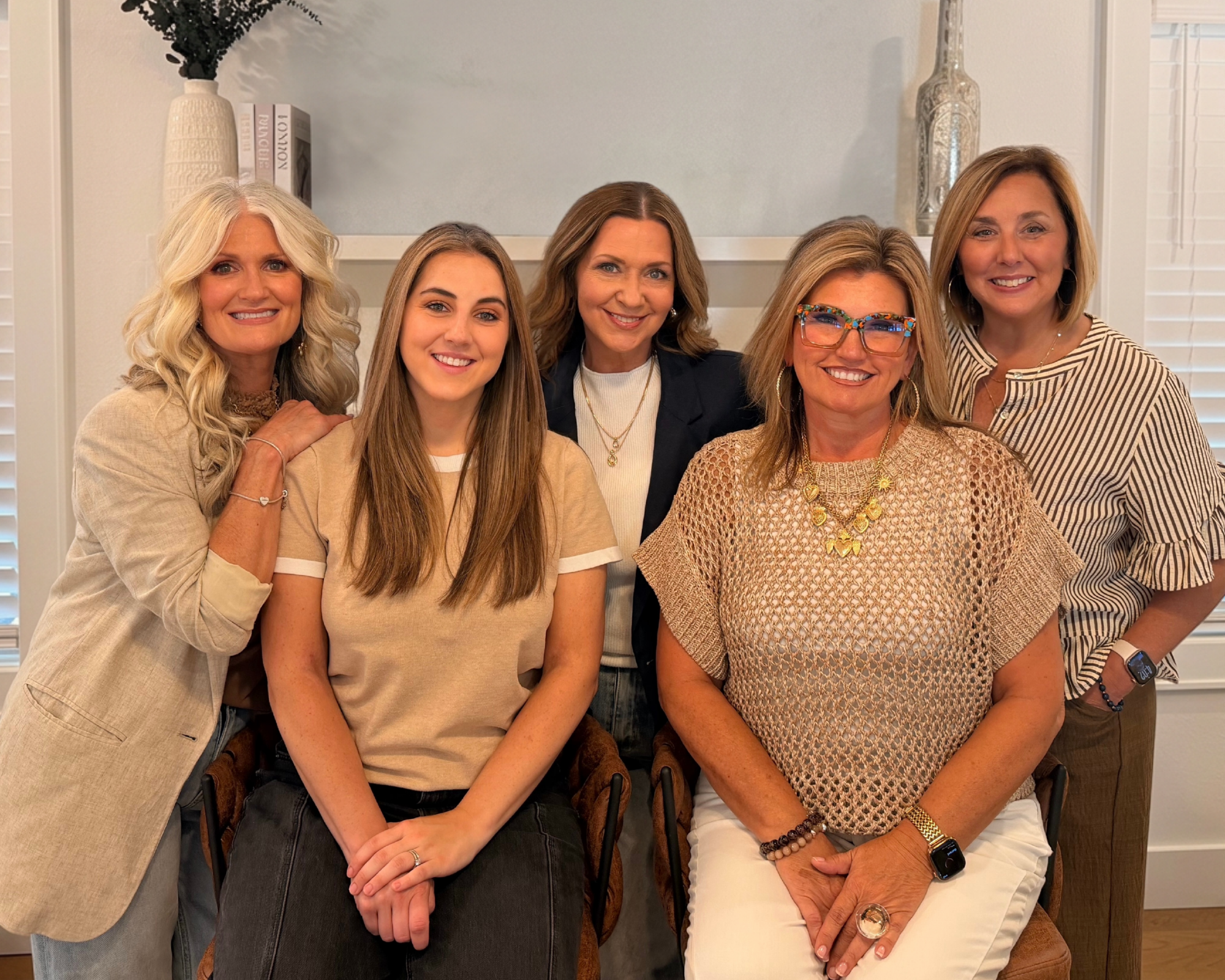 Group of five women smiling and posing together in an indoor setting, with their arms around each other.