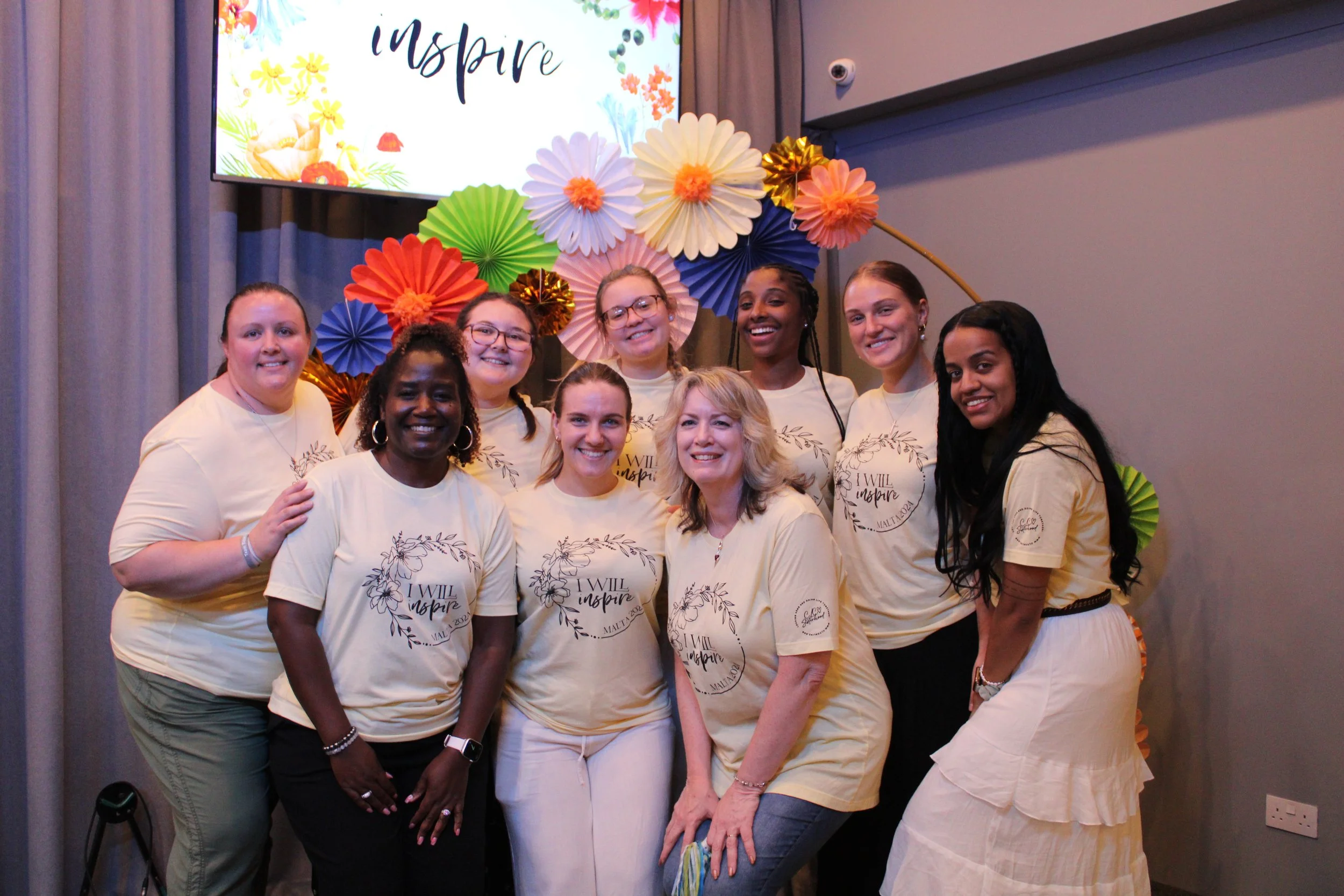 A diverse group of nine women standing together in front of a colorful backdrop of paper flowers and a screen displaying the word 'inspire' in stylized font. They are smiling and wearing matching cream-colored t-shirts with inspirational writing on them.