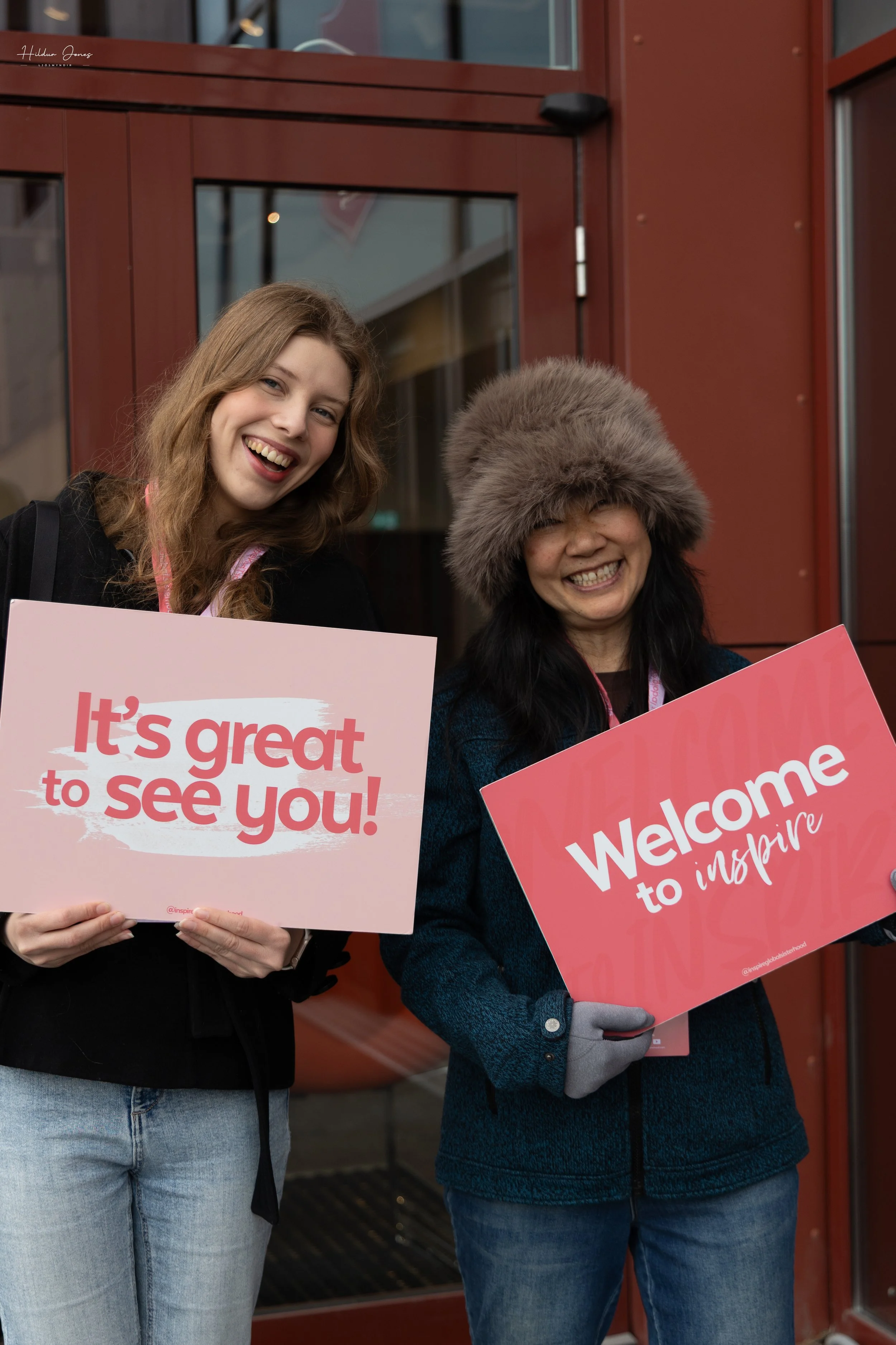 Two women standing outside a building holding signs that say 'It's great to see you!' and 'Welcome to inspire' and smiling.