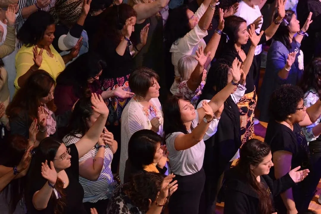 A diverse group of women at a religious or spiritual gathering, with many raising their hands in prayer or worship.