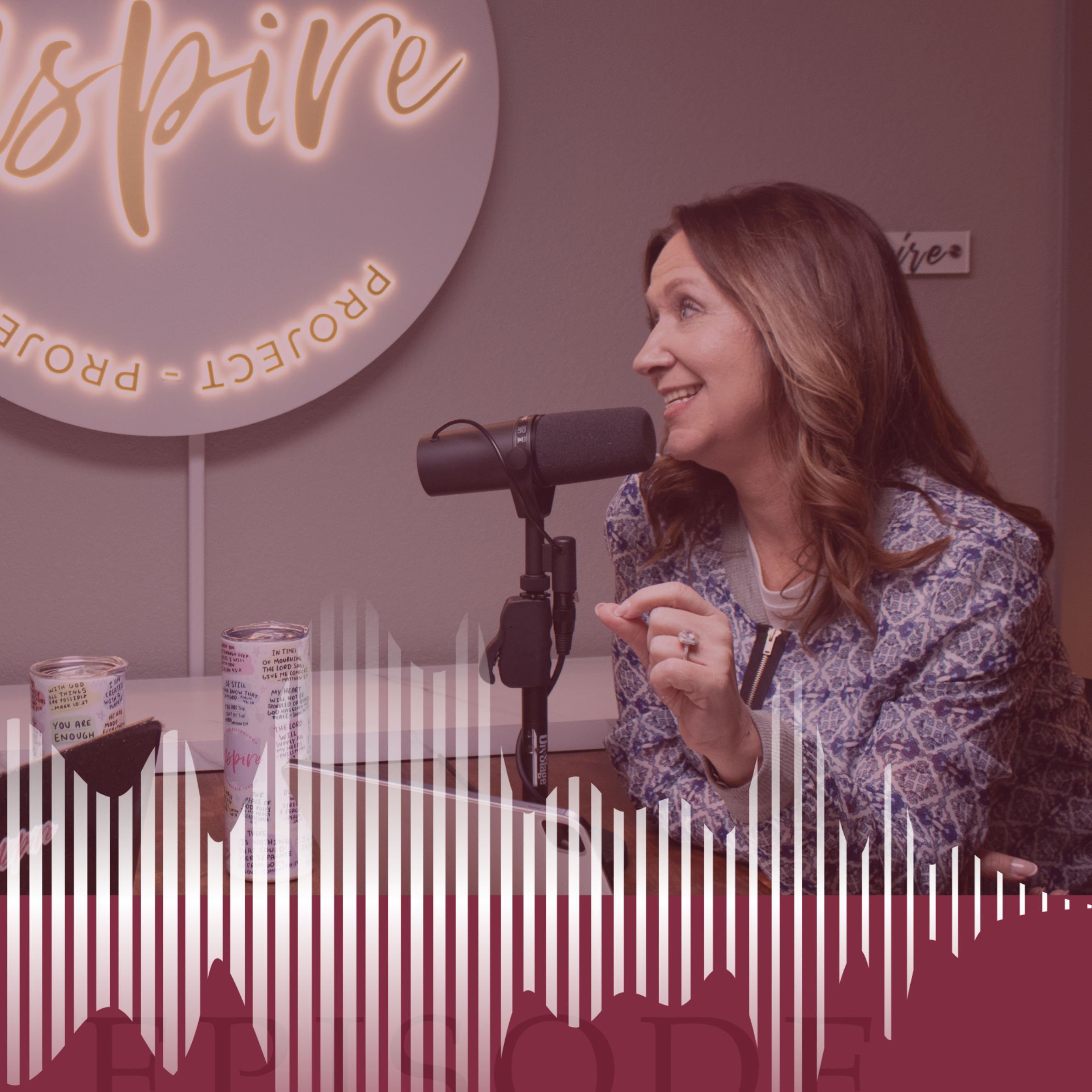 A woman with long brown hair smiling and speaking into a microphone during a podcast recording, with a neon sign in the background that says 'inspire' and a table with candles.