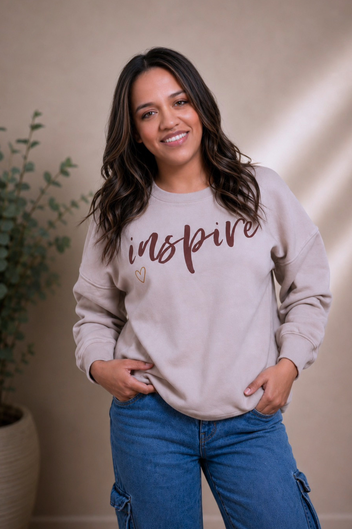 A woman with wavy brown hair, smiling, wearing a beige sweatshirt with the word 'inspire' written on it, and blue jeans, standing indoors with a potted plant in the background.