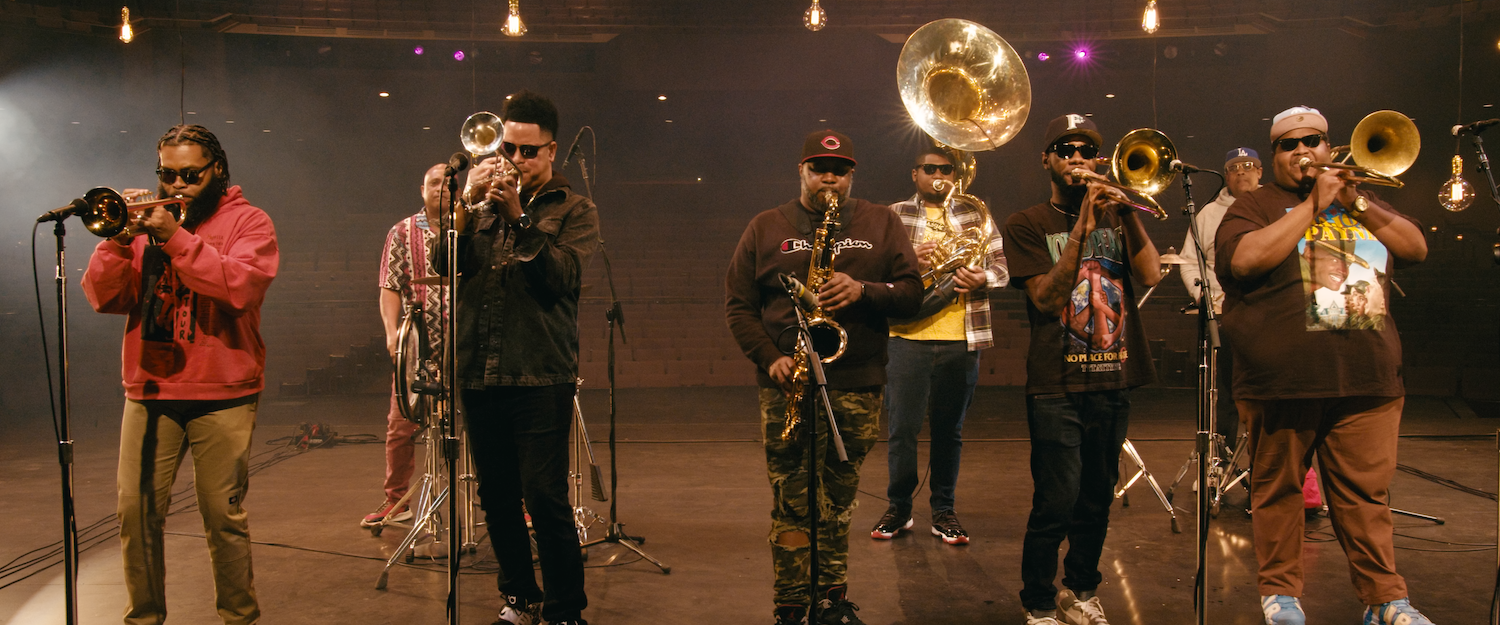 A band of seven musicians playing brass and wind instruments on a stage with dim lighting and hanging light bulbs.